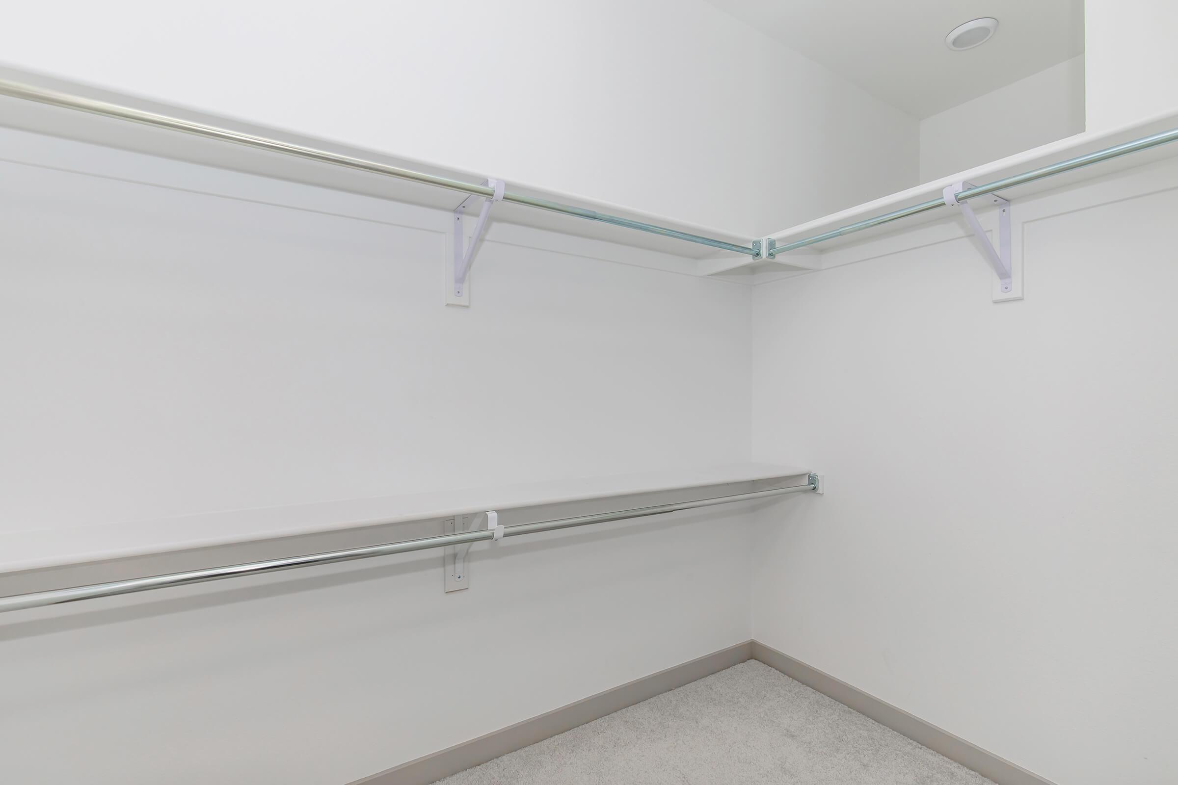 A spacious, empty closet featuring two metal hanging rods and a lower shelf, all arranged against white walls. The closet floor is carpeted, creating a clean and minimalistic look.