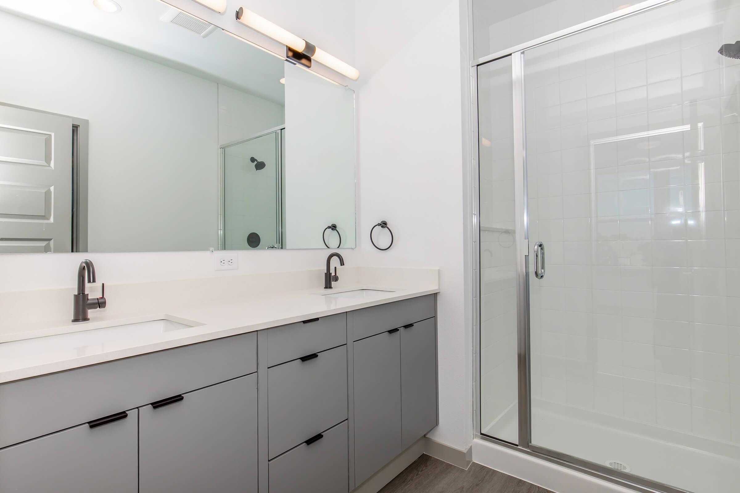 Modern bathroom featuring a double vanity with gray cabinetry, two sinks, and sleek black faucets. A large mirror spans the length of the vanity. To the right, there is a glass-enclosed shower with a tiled interior. The overall design is contemporary and minimalistic, with neutral colors and clean lines.