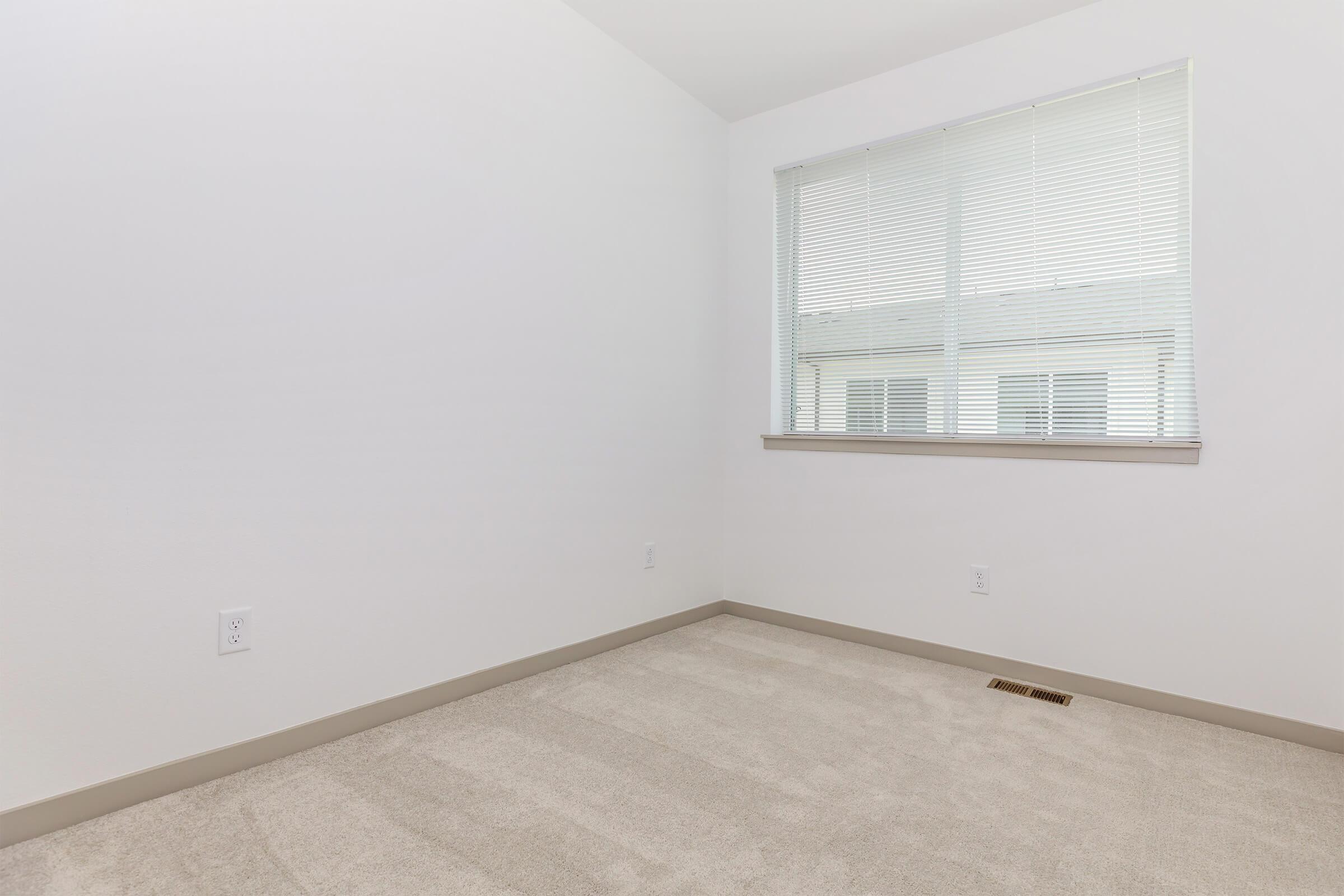 Empty room with light-colored carpet and a large window covered by blinds. The walls are painted white, and there are no furnishings or decorations present. The space appears clean and bright, suggesting a minimalistic design.