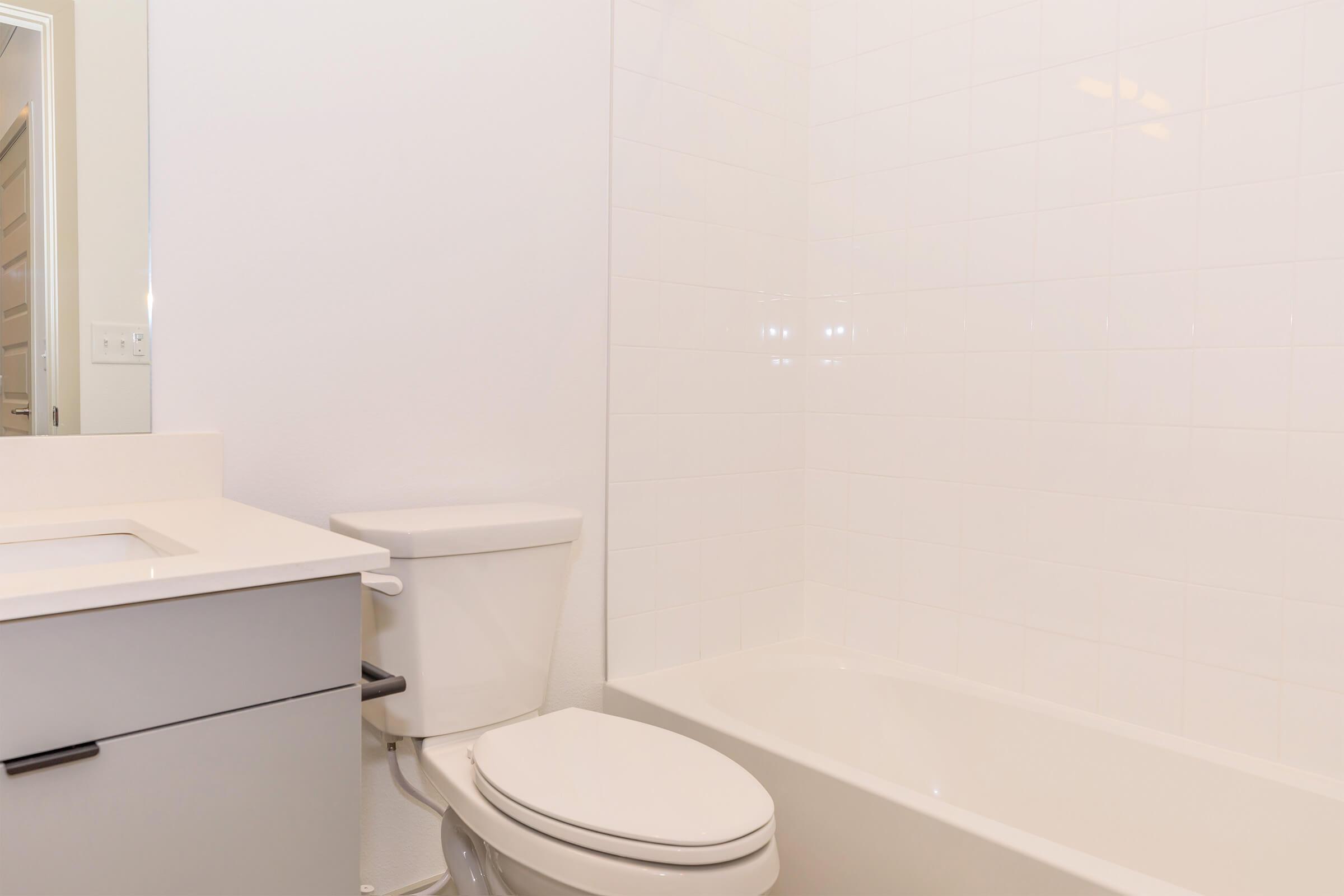 A clean, modern bathroom featuring a white tiled wall, a bathtub with a shower, a white toilet, and a sleek gray vanity with a sink. The overall design is minimalistic and bright, creating a fresh and open atmosphere.