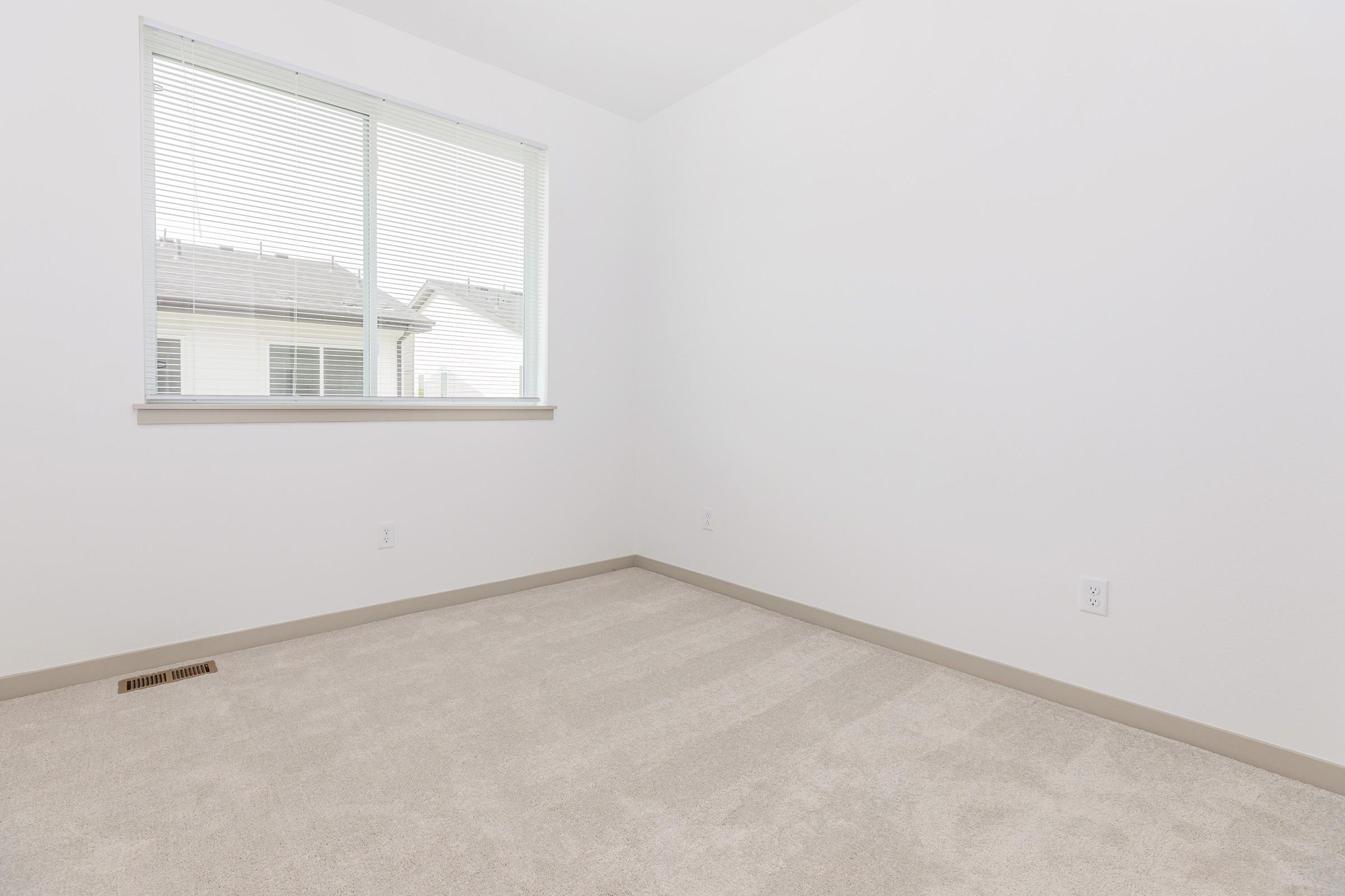 Empty room with light beige carpet and white walls. A large window with thin blinds allows natural light to enter the space. There are no furnishings or decorations, creating a minimalistic and clean atmosphere. The room has a neutral color scheme and a small vent on the floor.
