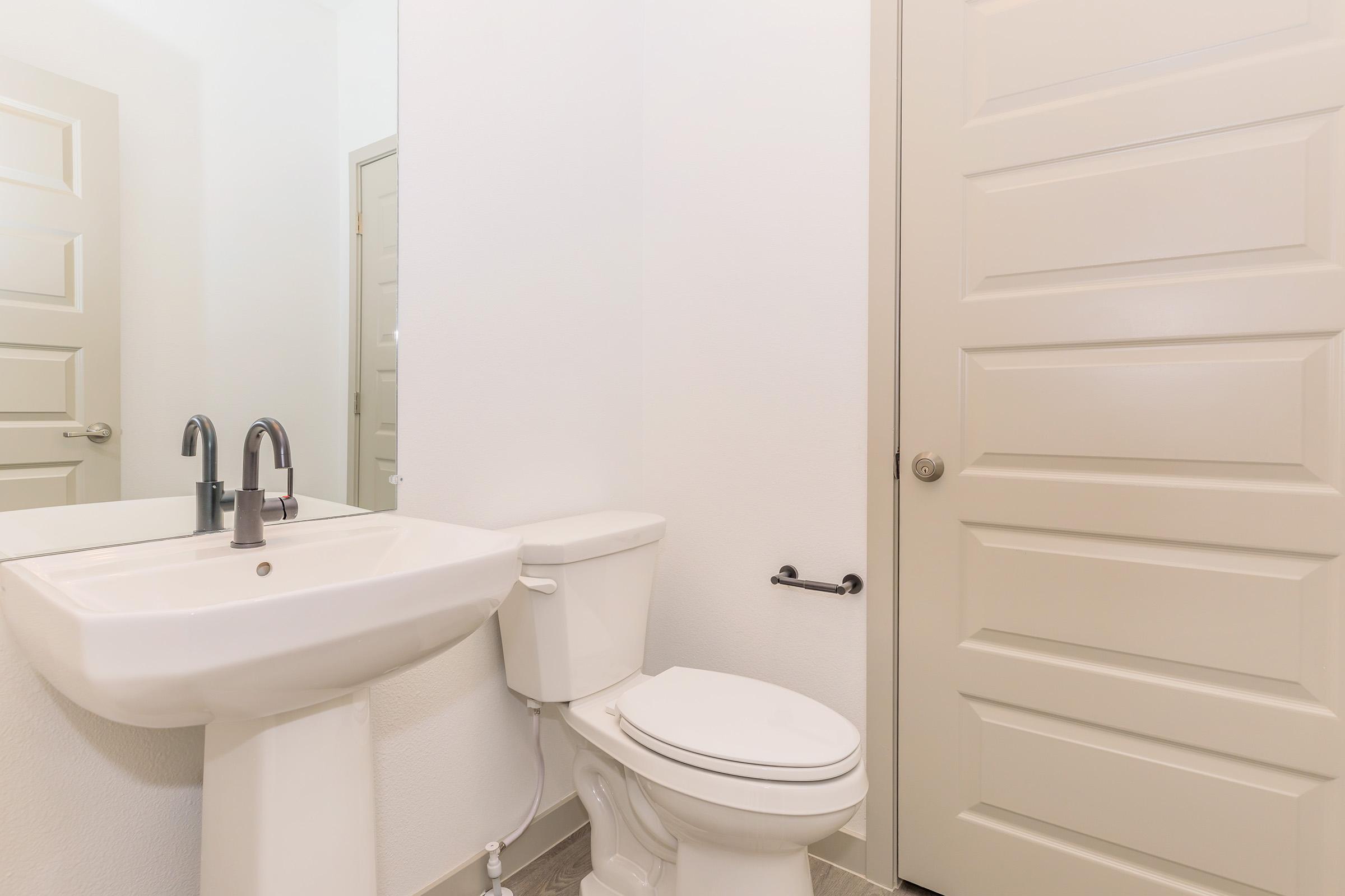 A clean bathroom featuring a white sink with modern faucets, a white toilet, and a closed beige door. The walls are painted white, and there is a mirror above the sink, reflecting the space. Neutral-colored tiles are visible on the floor.