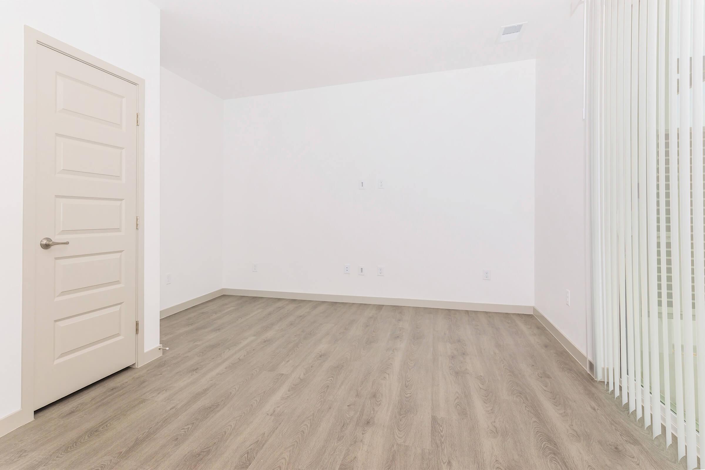 A bright, empty room featuring white walls and light wood-like flooring. The room includes a closed door on the left and vertical blinds on the right, with no furnishings or decorations present. Natural light may be coming through the blinds, creating a spacious and clean appearance.