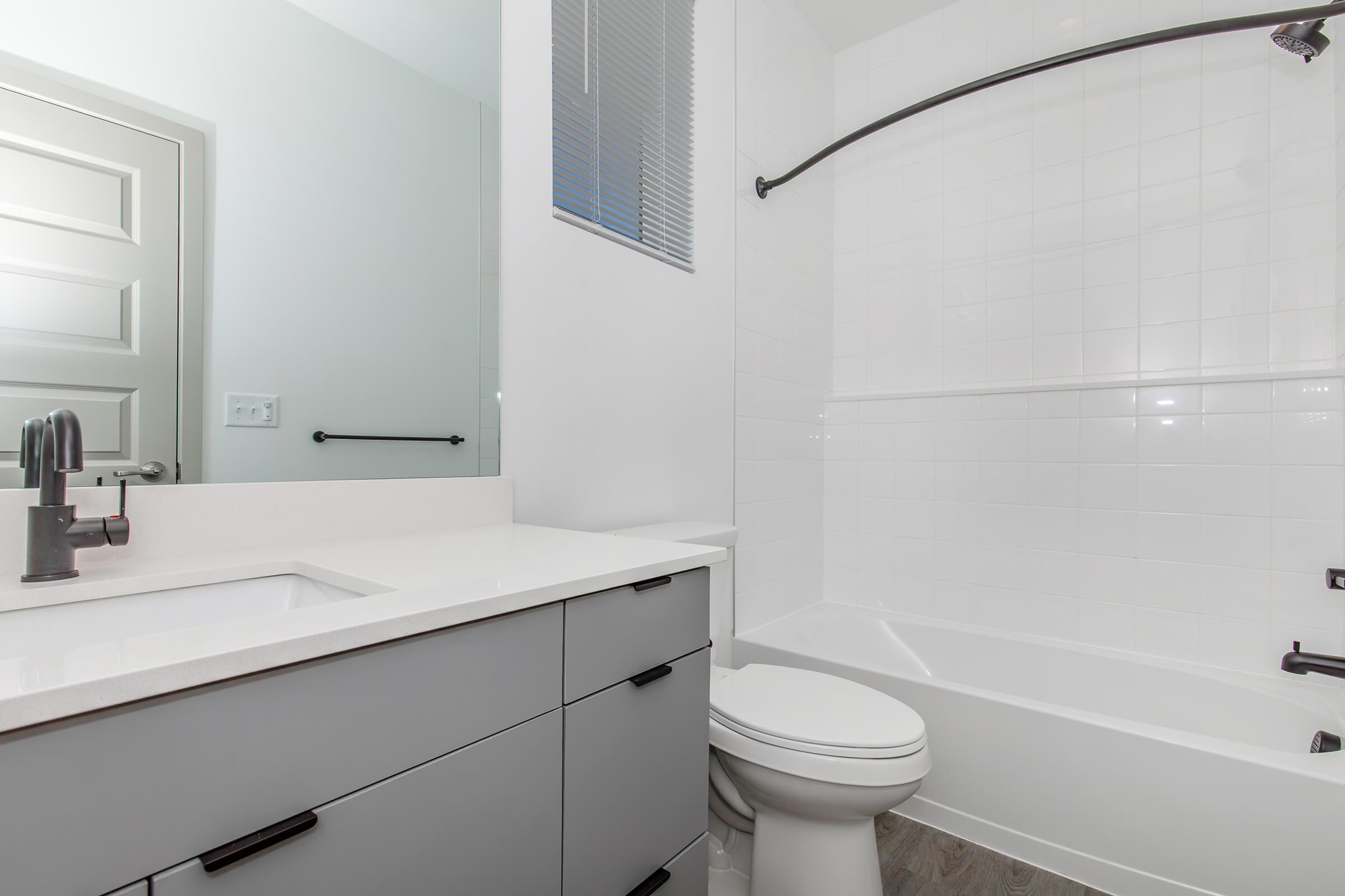 Modern bathroom featuring a white tiled shower/tub combination, a sleek vanity with a solid surface countertop, and a stylish faucet. The space includes a large mirror above the sink and a door leading to another area, with light-colored walls and gray accents.