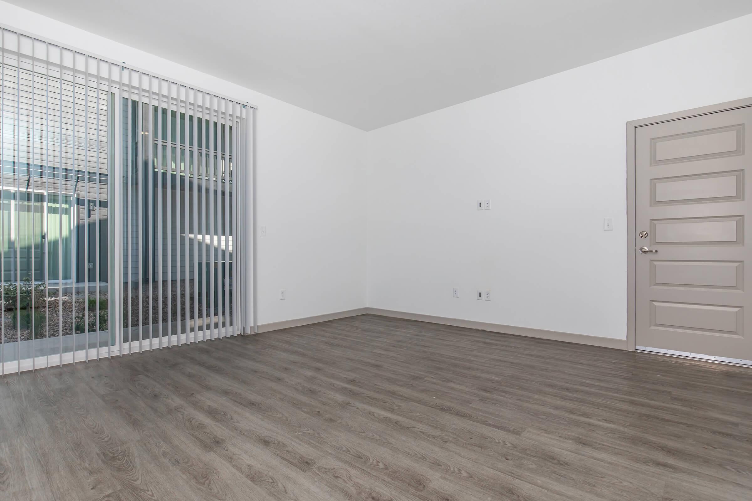 Empty room with light-colored walls and laminate flooring. A large window with vertical blinds allows natural light to enter. There is a door on the right side leading outside, and no furniture or decorations are present, creating a spacious and minimalistic atmosphere.
