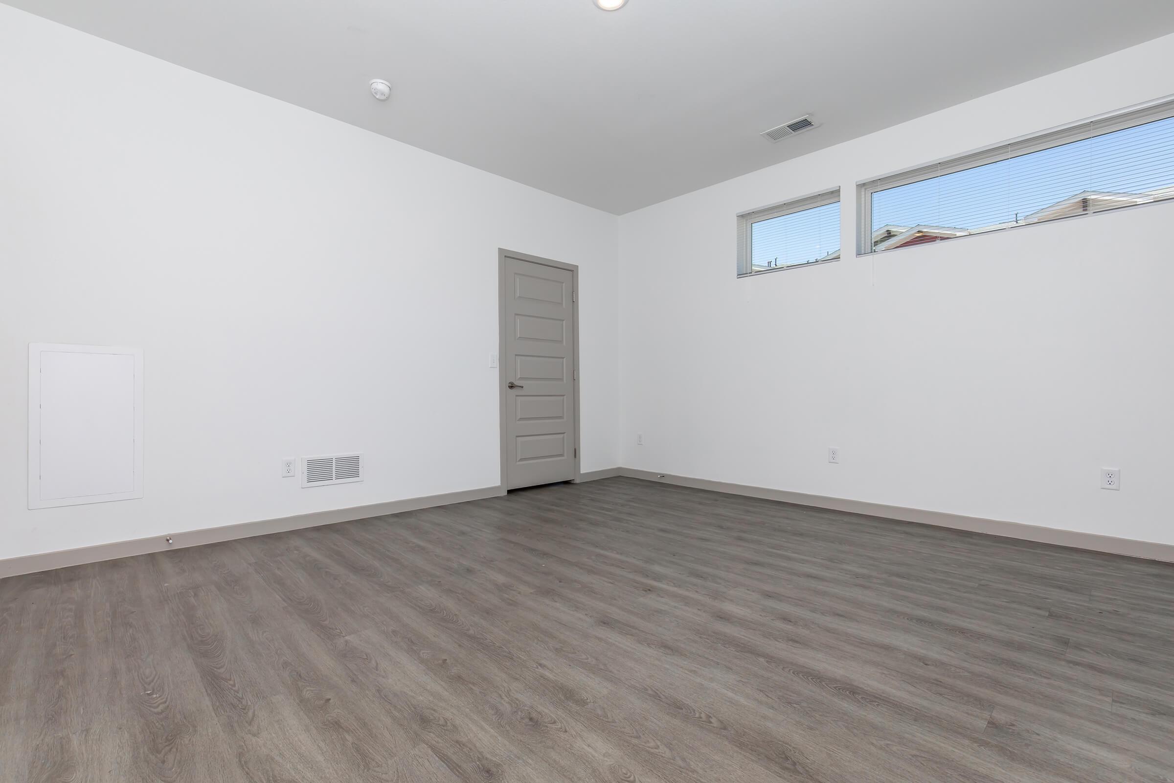 Empty interior of a room with light-colored walls and a door on the side. The floor is made of light wood or laminate. There are two small windows letting in natural light. A vent is visible on the wall, and a small panel is located near the floor. The room appears modern and minimalist.