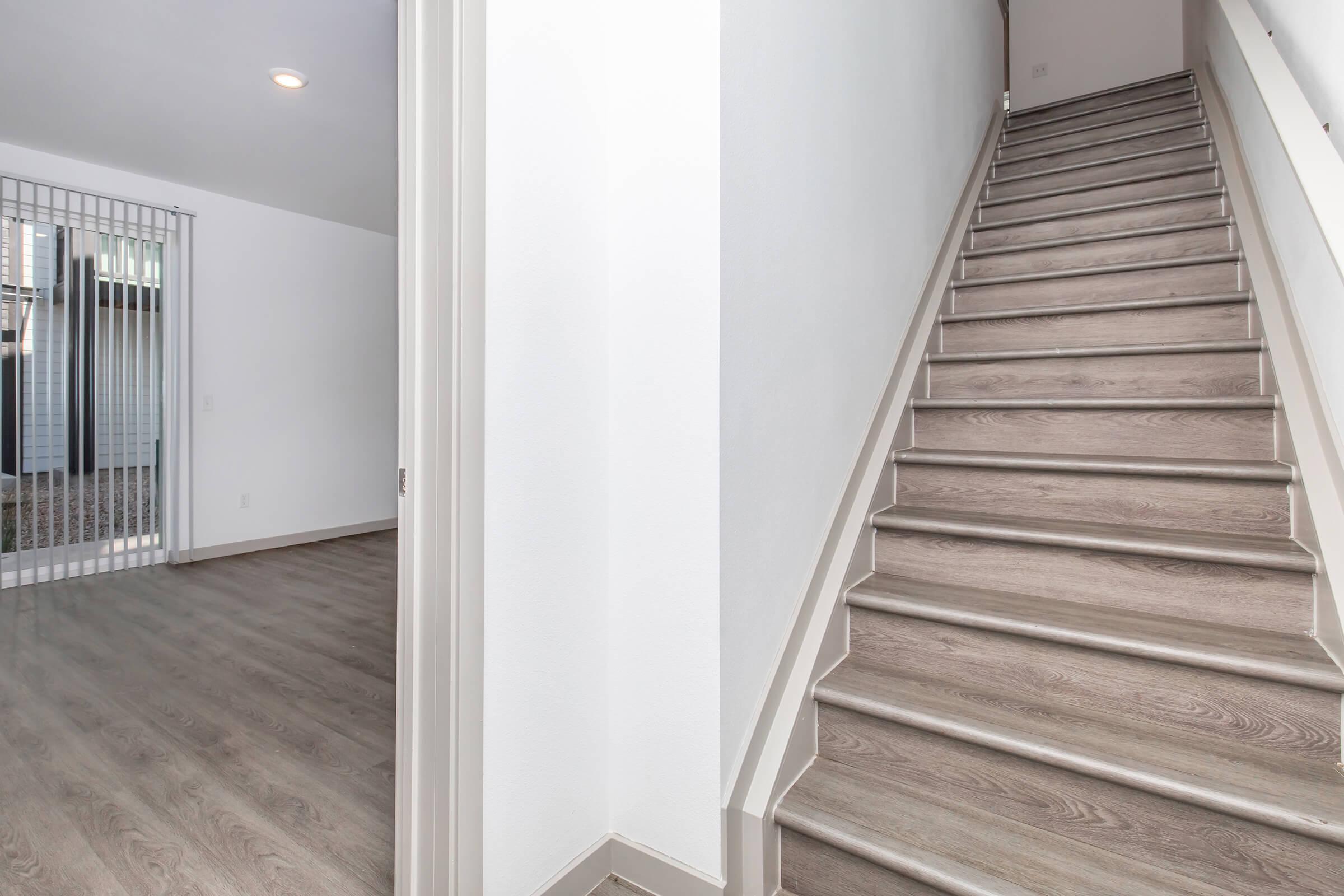 Interior view of a modern home featuring a staircase with wooden steps leading to an upper level. To the left, there is a doorway leading to a room with a large window and a patterned carpet. The walls are painted white, and the flooring is light-colored wood.