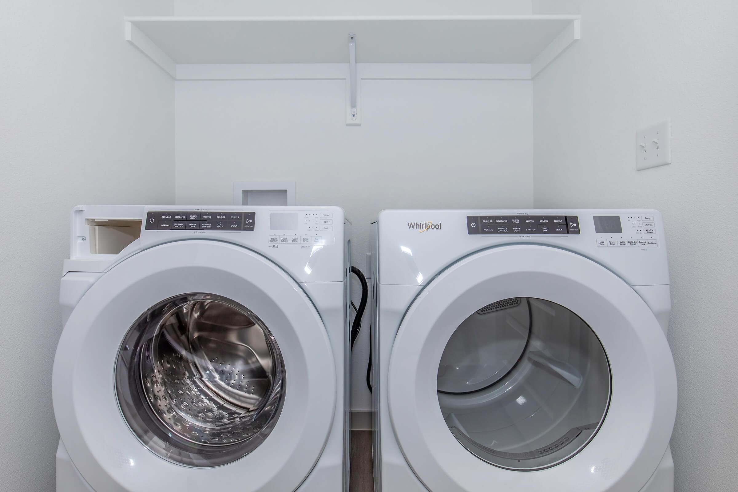 A modern laundry room featuring a pair of white Whirlpool washing machines side by side. Above them is a simple shelf for storage, with a clean, minimalist wall in the background. The machines are front-loading, showcasing their sleek design and control panels.