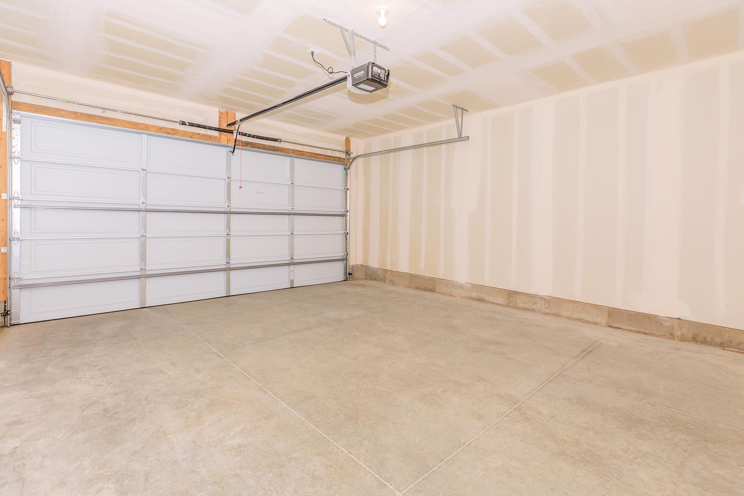 A spacious, empty garage with a concrete floor and unfinished walls. The garage features a large, gray overhead door on one side, and a ceiling with exposed beams and lights. The space is well-lit, providing ample room for parking or storage.