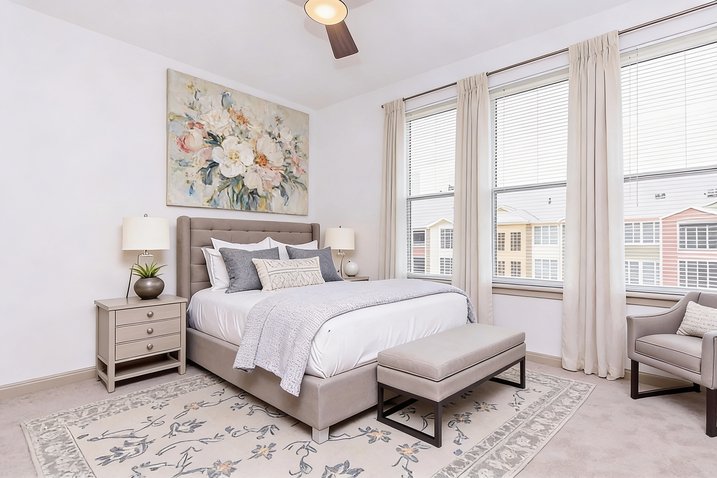 A stylish bedroom featuring a gray upholstered bed with decorative pillows, a light gray throw blanket, and a matching ottoman. There are two bedside lamps on nightstands and a floral artwork above the bed. The large windows allow natural light, with sheer curtains and a view of colorful buildings outside.