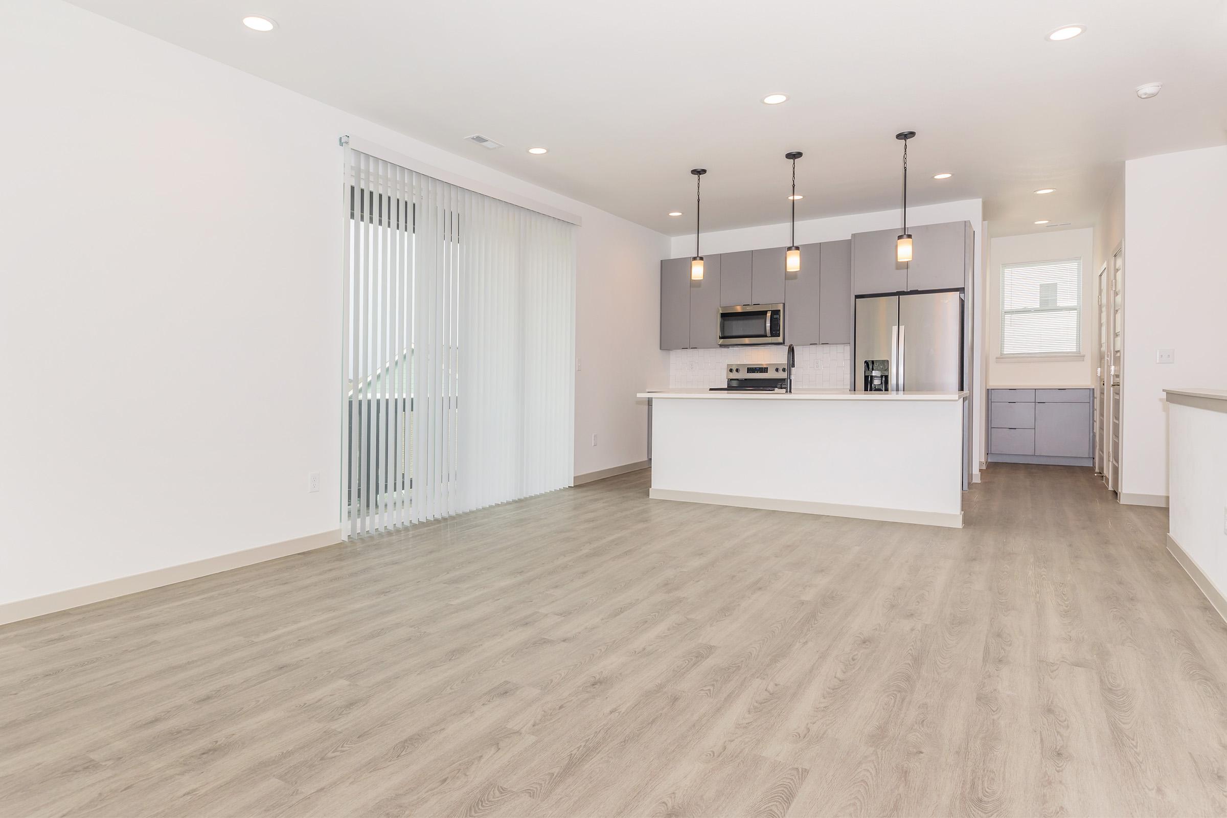 Modern open-concept living space featuring light wood flooring, a sleek kitchen with stainless steel appliances, and pendant lighting. Large windows provide natural light, and a sliding door leads to an outdoor area. The walls are painted white, creating a bright and airy atmosphere.