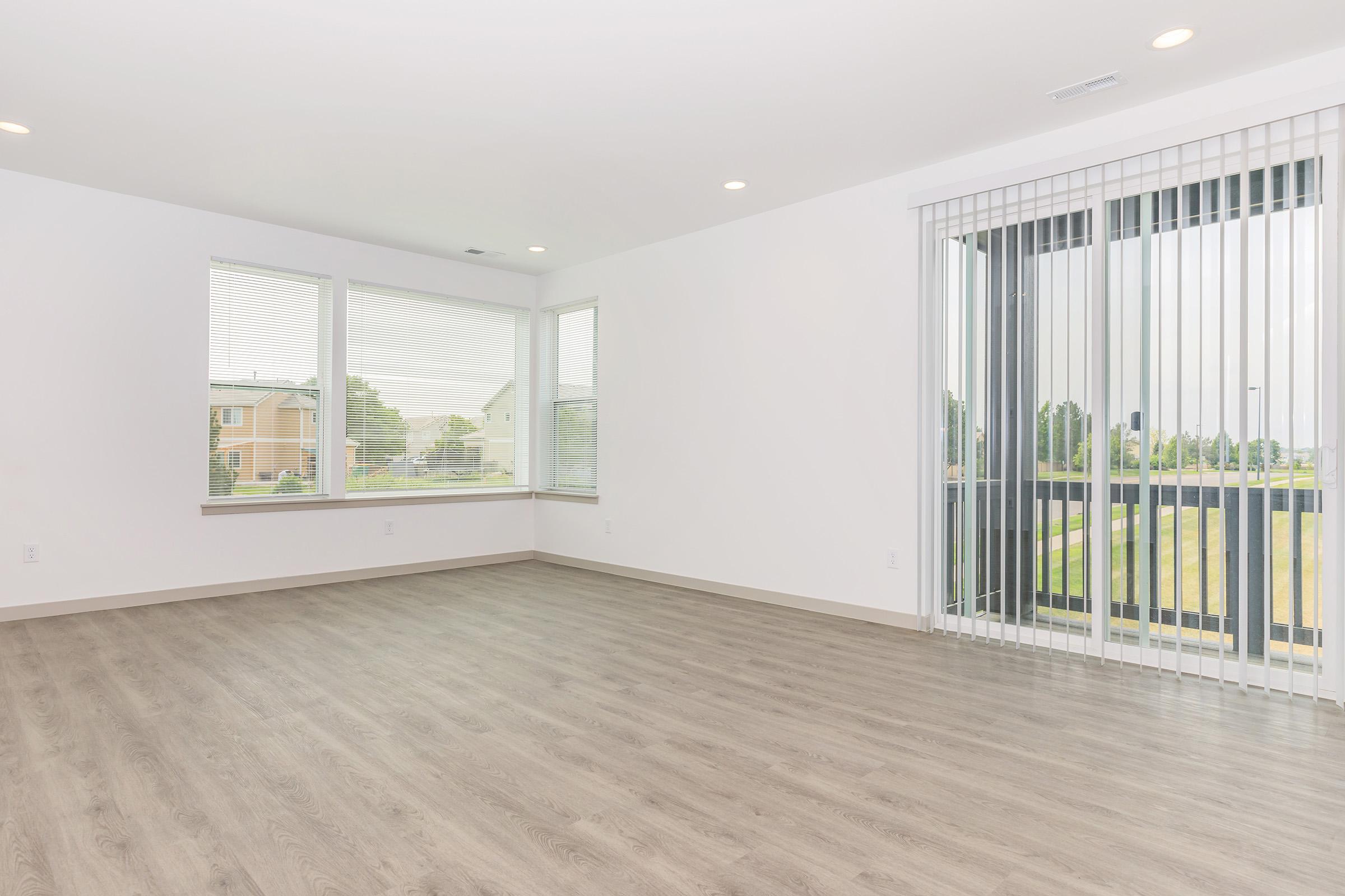 Spacious interior of a modern room with large windows, allowing natural light to fill the space. Light-colored walls complement the wood-like flooring. Vertical blinds cover the windows. A view of greenery is visible outside. The room is empty, showcasing the potential for furniture arrangement.