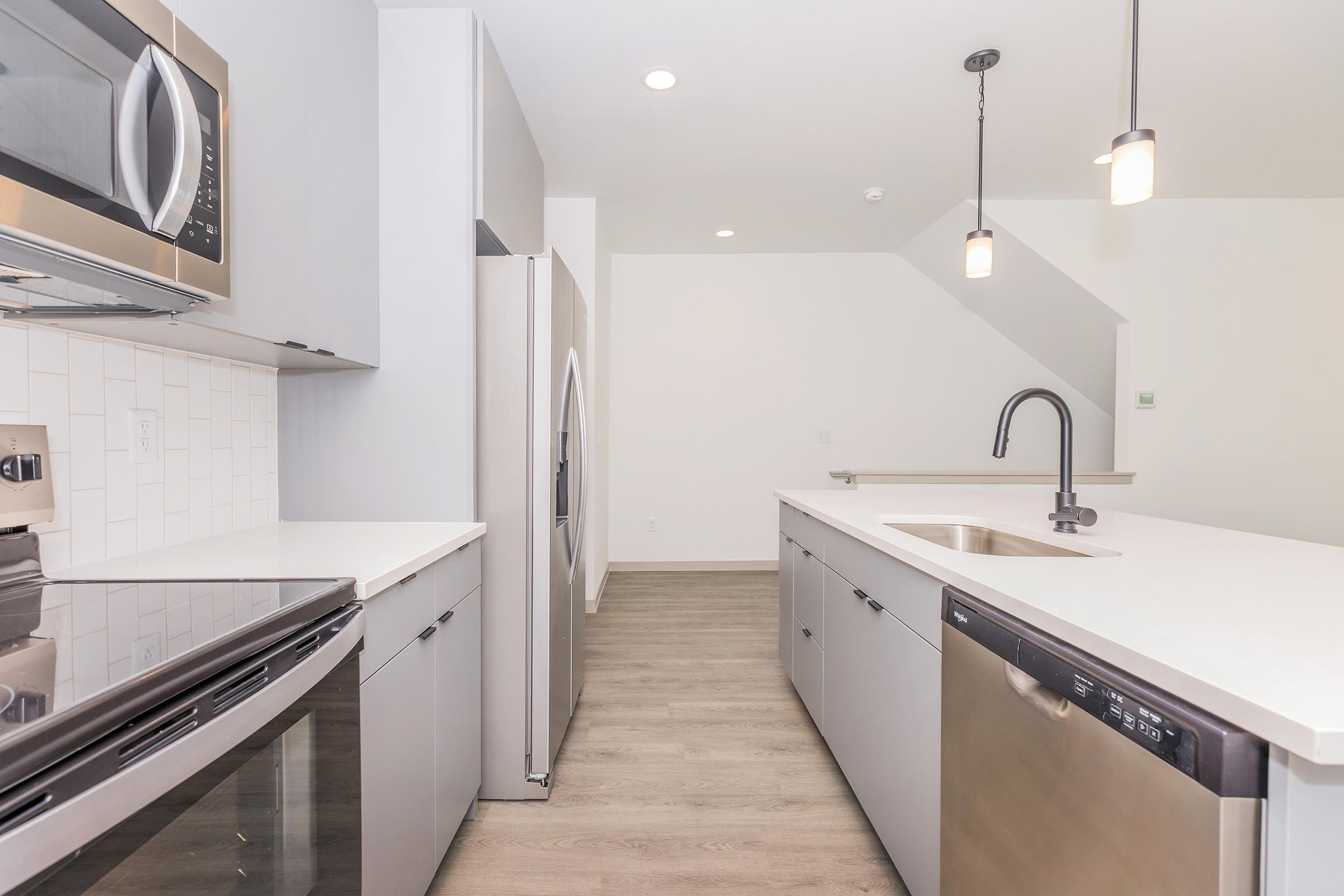 Modern kitchen featuring sleek gray cabinetry, stainless steel appliances including a microwave, oven, refrigerator, and dishwasher. A countertop with a stylish sink and a single pendant light illuminate the space. Light wood flooring enhances the contemporary design. The layout is open and airy.