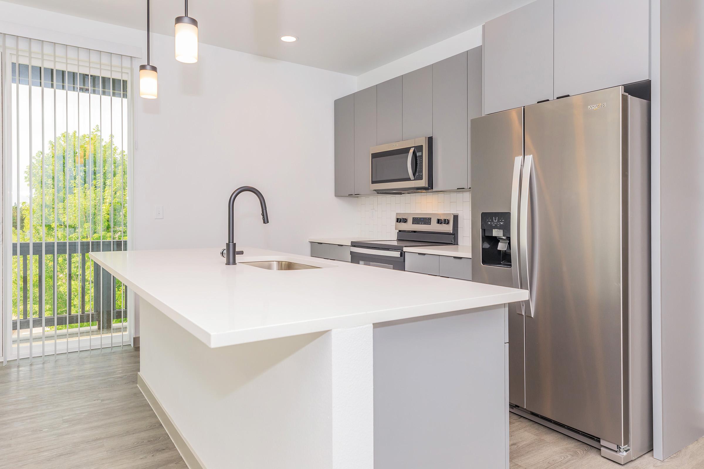 Modern kitchen featuring gray cabinetry, stainless steel appliances including a refrigerator and microwave, a white countertop, and a sleek black faucet. A large window with vertical blinds allows natural light, enhancing the contemporary design. The flooring is light wood, complementing the overall aesthetic.