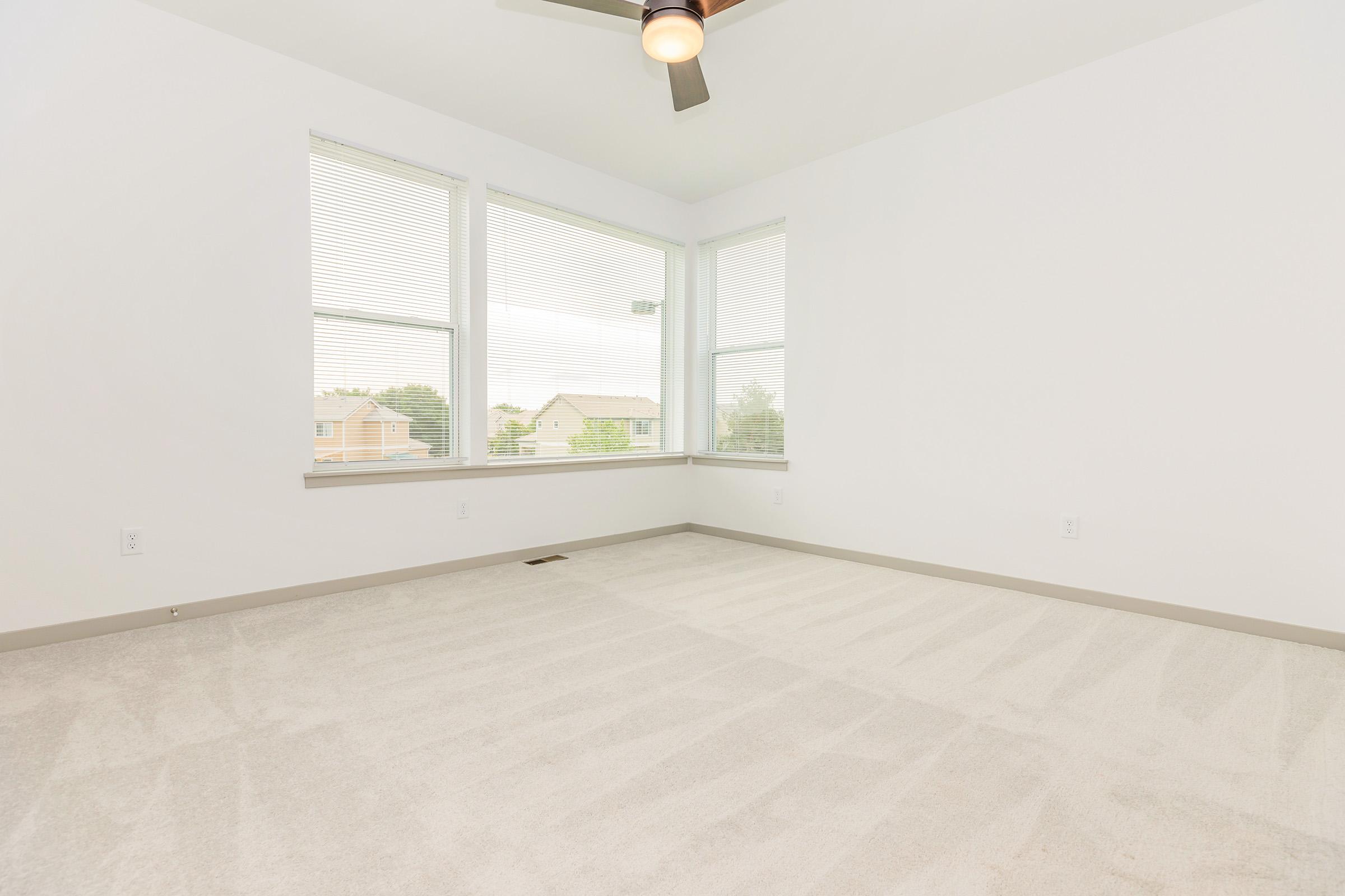 A spacious, empty room with light-colored walls and a soft carpet. It features three large windows that allow natural light to fill the space. A ceiling fan is visible, and the room has a clean, minimalist appearance, making it suitable for various uses.
