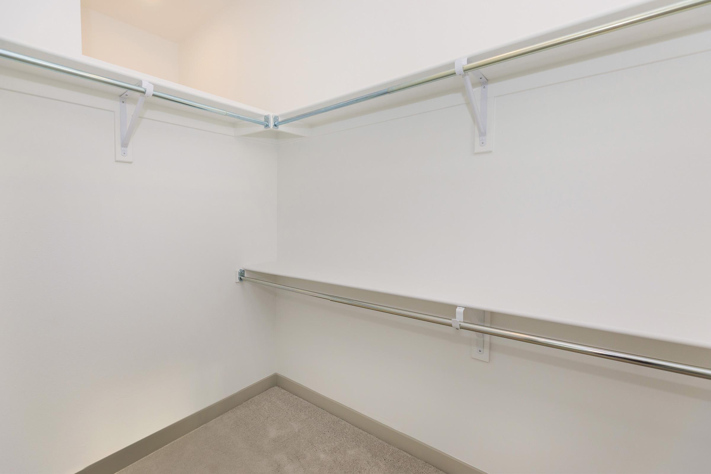 A clean, empty closet with white walls and shelving. The closet features two horizontal rods for hanging clothes, with a light beige carpeted floor. The space is well-lit and minimalistic, emphasizing its emptiness. Ideal for organizing personal items.