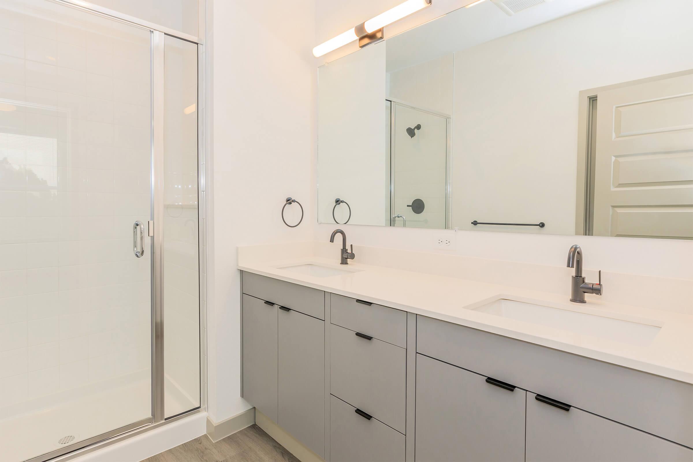 Modern bathroom featuring a double vanity with two sinks, sleek gray cabinets, and a large mirror. There's a glass shower stall with a rainfall showerhead and a light neutral tile floor, creating a spacious and contemporary feel.
