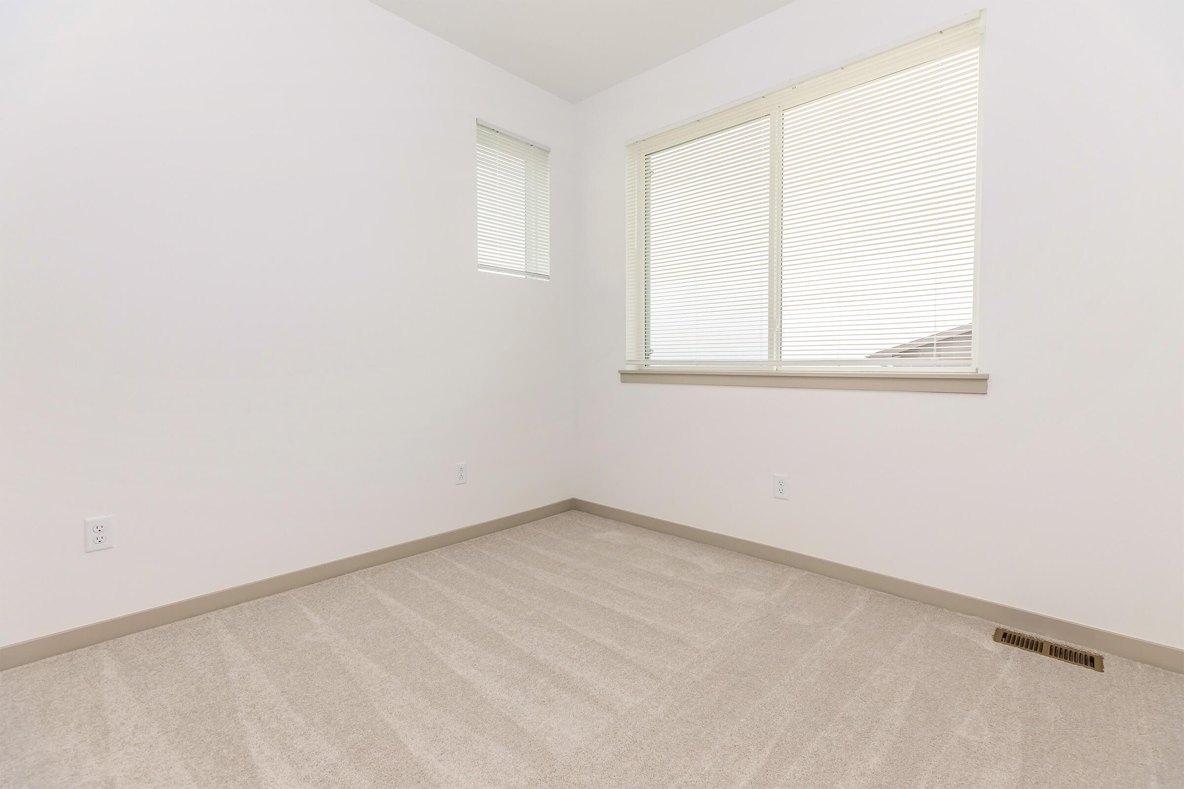 Empty room with light-colored walls and beige carpet. A large window with horizontal blinds allows natural light to enter, casting soft shadows on the floor. There is a small, square window high on one wall. The space appears clean and uncluttered, suitable for various uses or decoration.