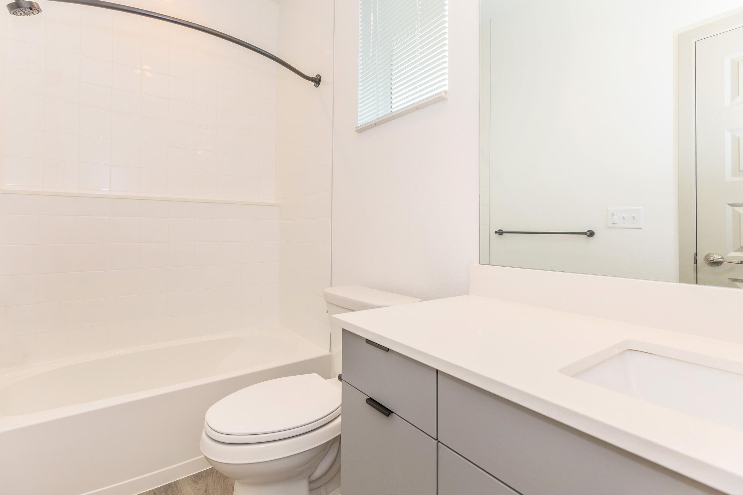 A modern bathroom featuring a white tiled bathtub with a shower, a sleek white countertop with a sink, and a stylish gray cabinet. The space is bright, with natural light and minimalistic decor, showcasing a clean and contemporary design.