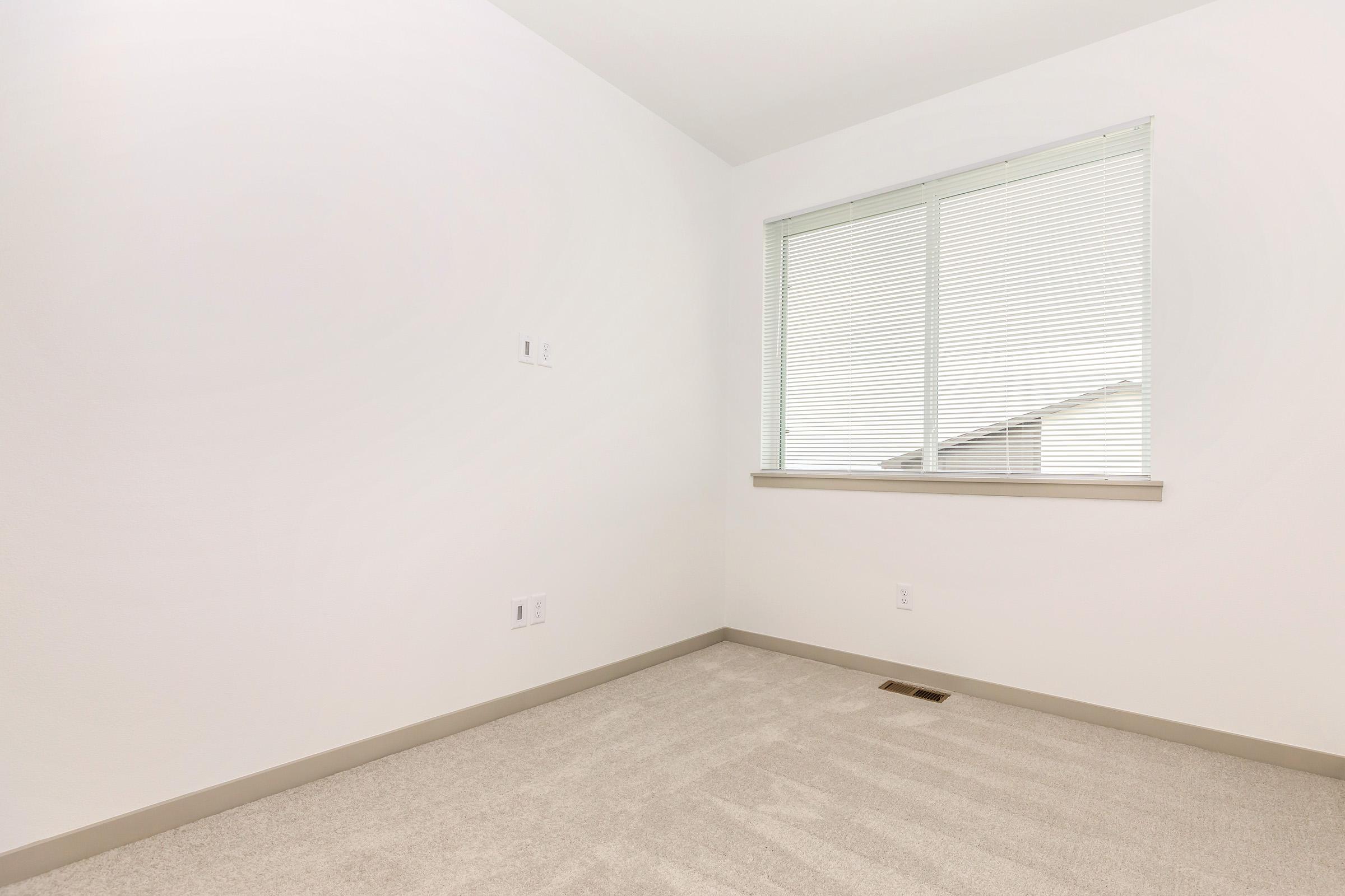 Empty room with white walls and a window featuring blinds, allowing natural light to enter. The carpet is light-colored and there are no furnishings or decorations in the space. A small section of the wall has electrical outlets.