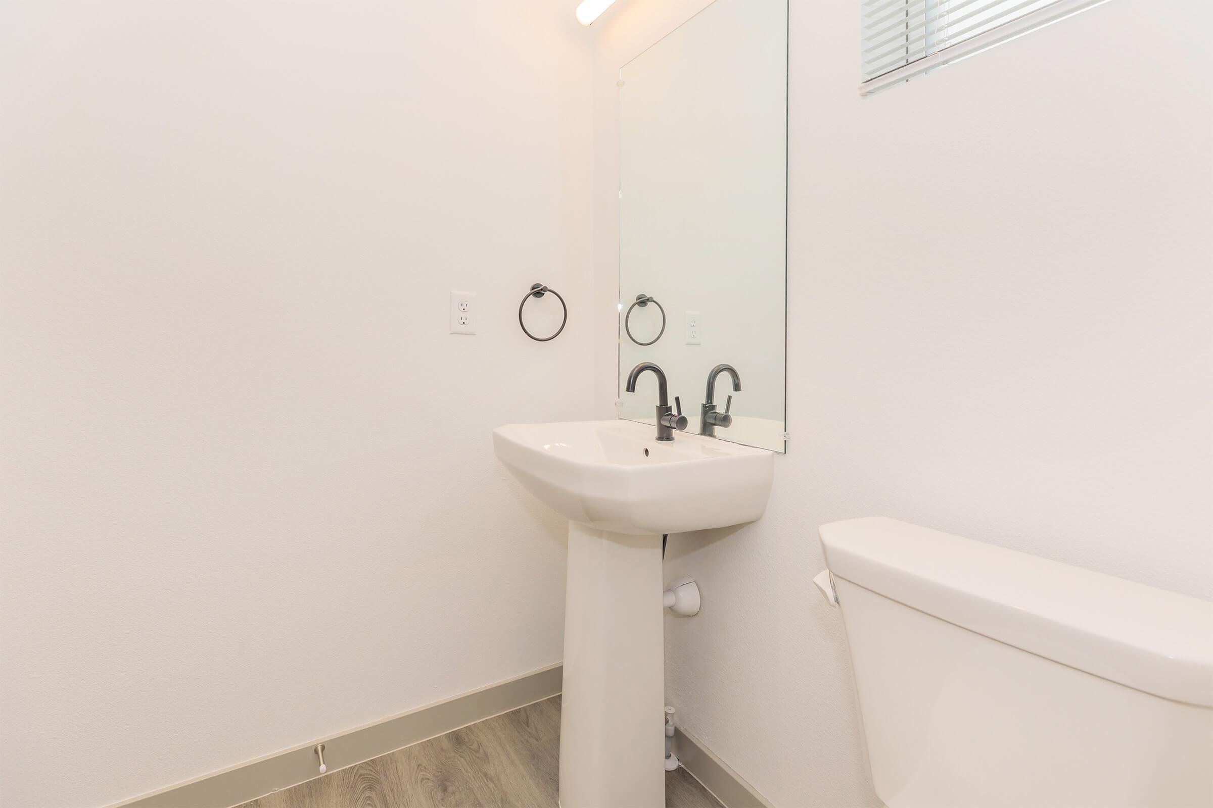 A clean and modern bathroom featuring a white pedestal sink with two faucets, a large wall mirror, a towel ring, and a white toilet. The walls are painted a light color, and there is a small window allowing natural light. The floor has a wood-like finish.