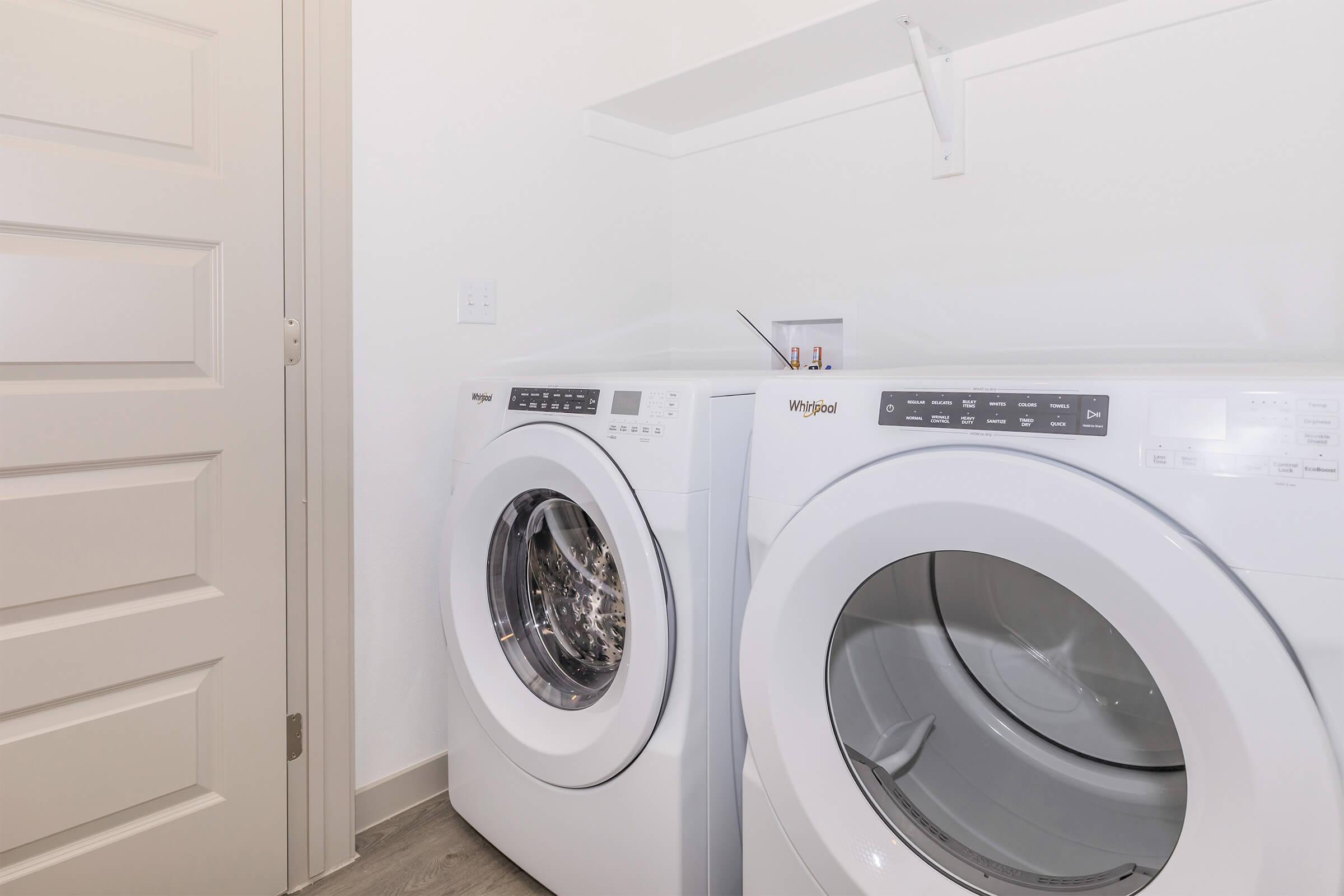 A clean laundry room featuring a pair of white Whirlpool washing machines stacked next to each other, with a light-colored door in the background and minimalist decor on the walls. The space appears bright and organized, ideal for household chores.