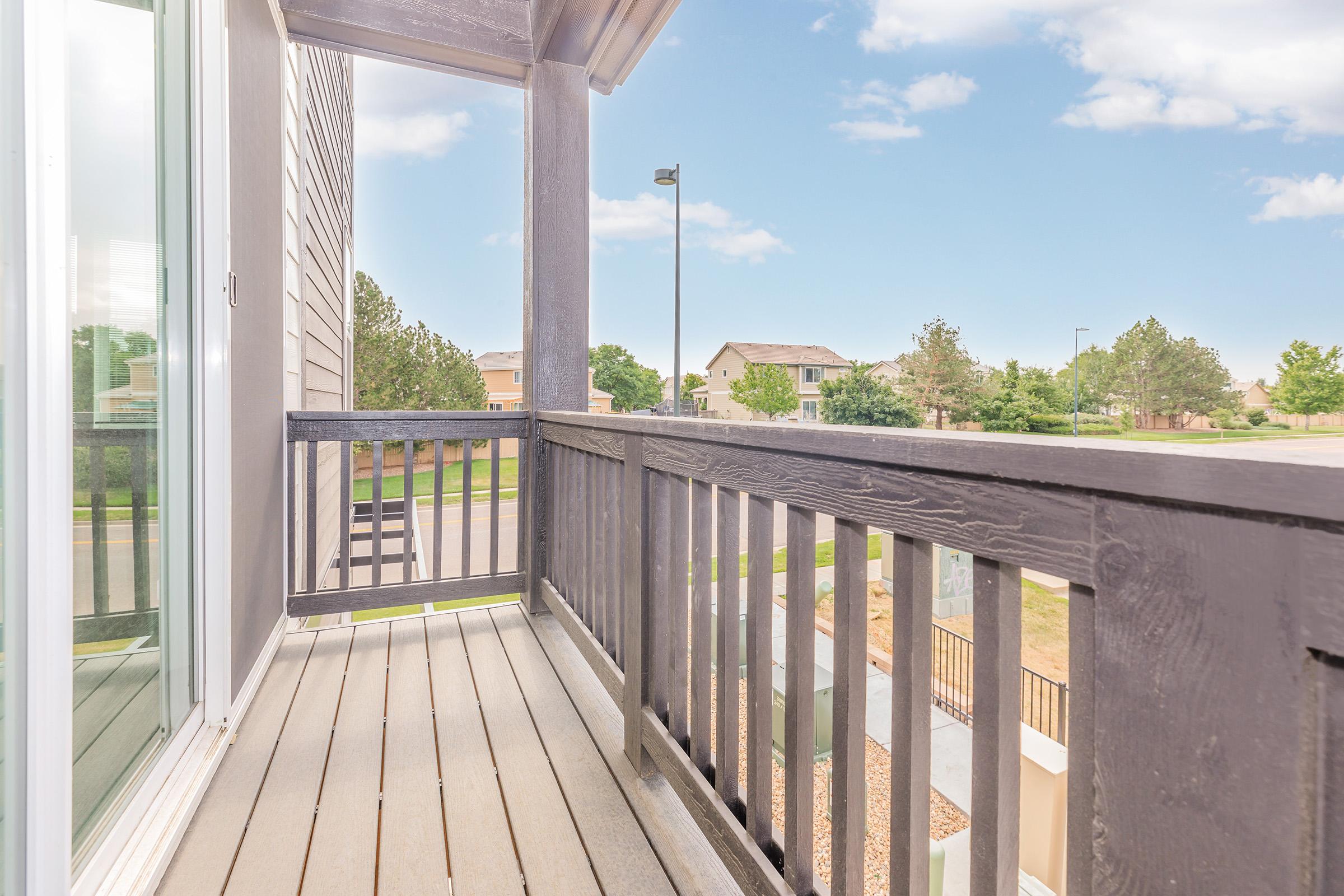 A wooden balcony with a railing, featuring a view of a neighborhood with houses and trees in the background under a partly cloudy blue sky. The scene is bright and inviting, with sunlight illuminating the space.