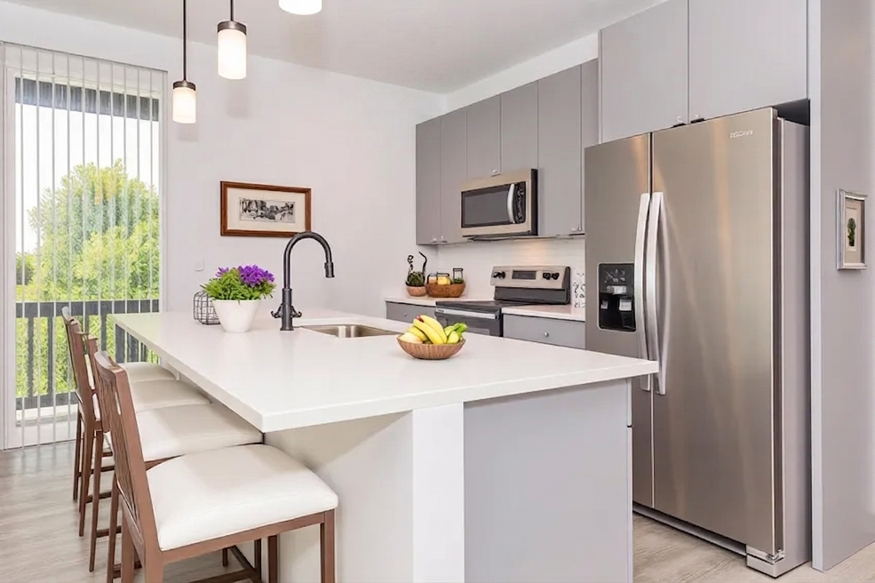 Modern kitchen featuring gray cabinetry, stainless steel appliances, and a large island with seating for four. A bowl of fruits sits on the counter, while a potted plant adds a touch of greenery. Natural light pours in through vertical blinds, creating a bright and inviting atmosphere.