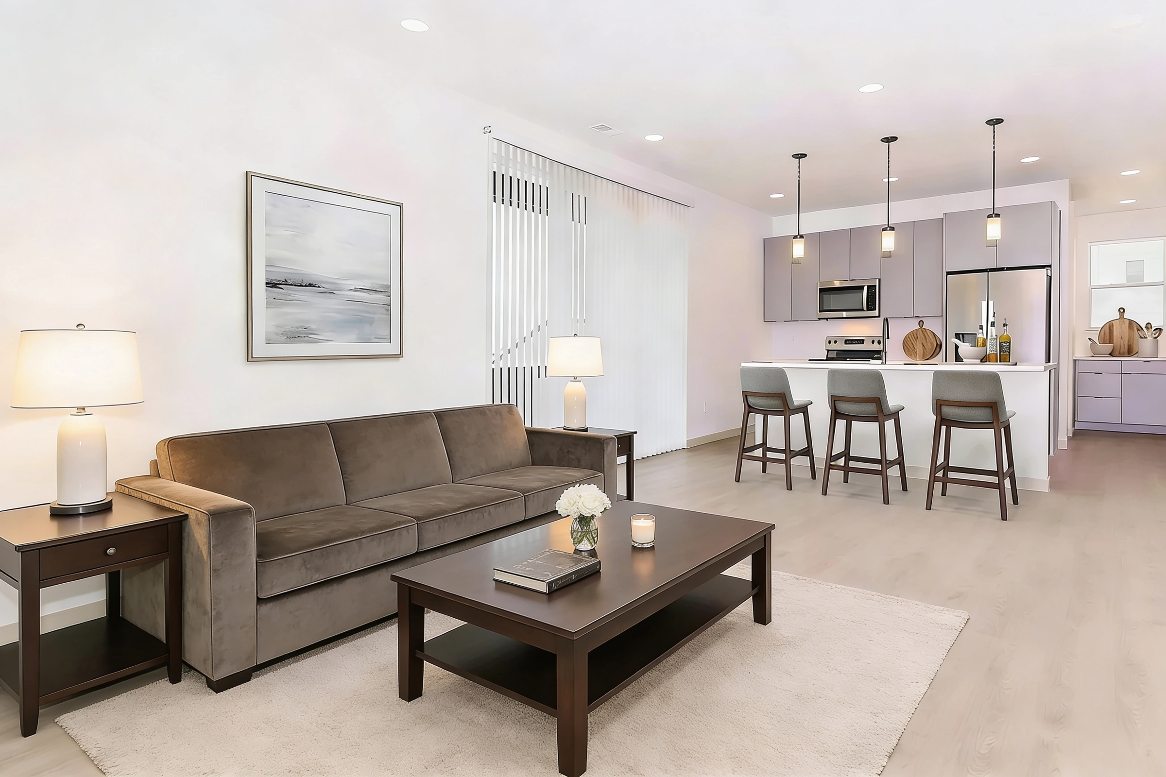 A modern living room featuring a brown sofa with a side table and a coffee table. In the background, a kitchen area with light purple cabinets, bar stools, and sleek appliances. Soft lighting from pendant lamps and lamps on side tables creates a warm ambiance. Light-colored flooring and a neutral rug complete the space.