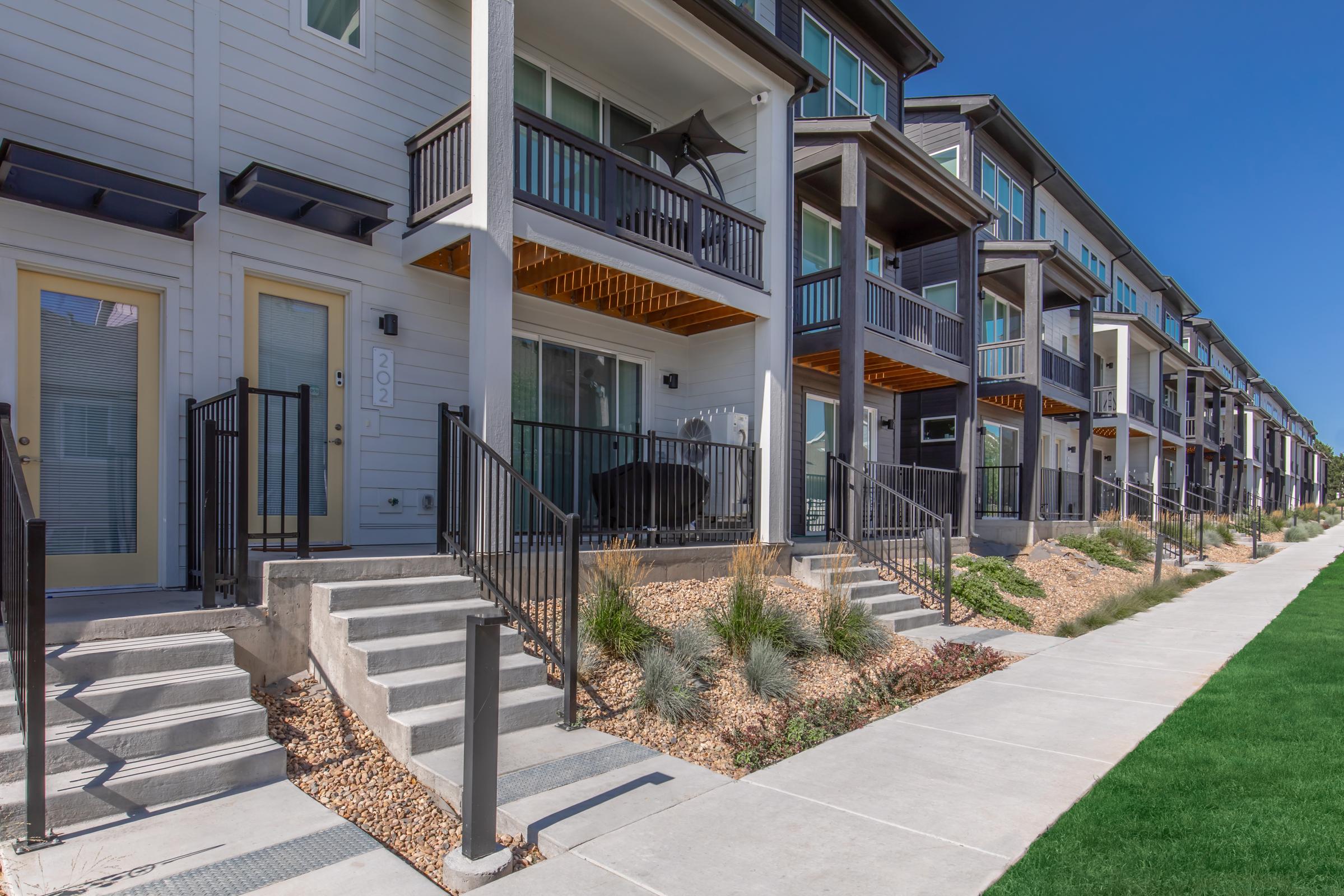 Row of modern townhomes with steps leading to individual entries. Each unit features balconies with black railings and large windows. The landscape includes low-maintenance plants and a neat walking path alongside green grass, under a clear blue sky.