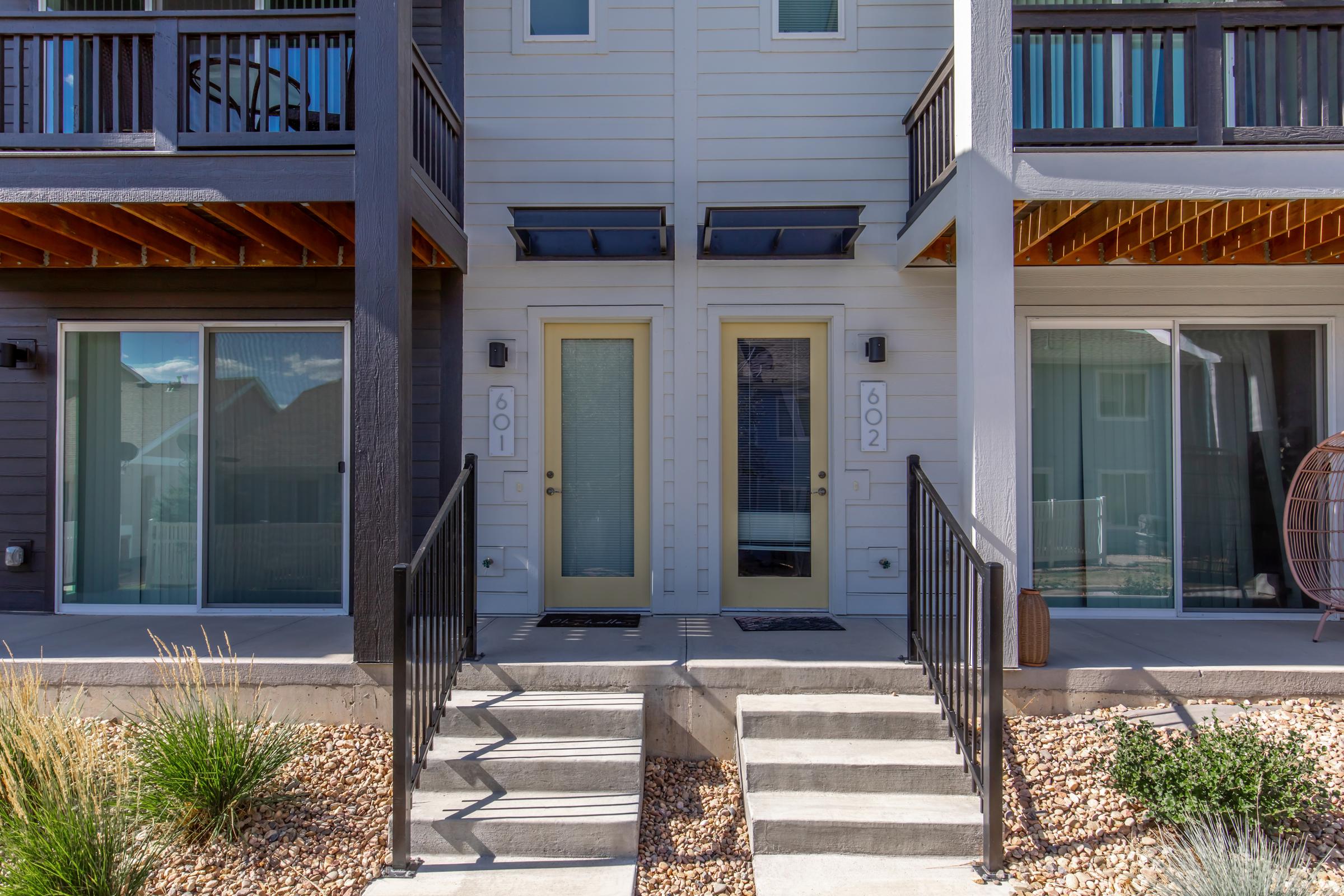 Two entry doors numbered 601 and 602, leading to residential units. The doors are framed by a porch area with wooden accents. Steps lead up to the doors, flanked by minimal landscaping featuring gravel and small plants. Large windows above the doors provide light, and a covered awning offers shelter.