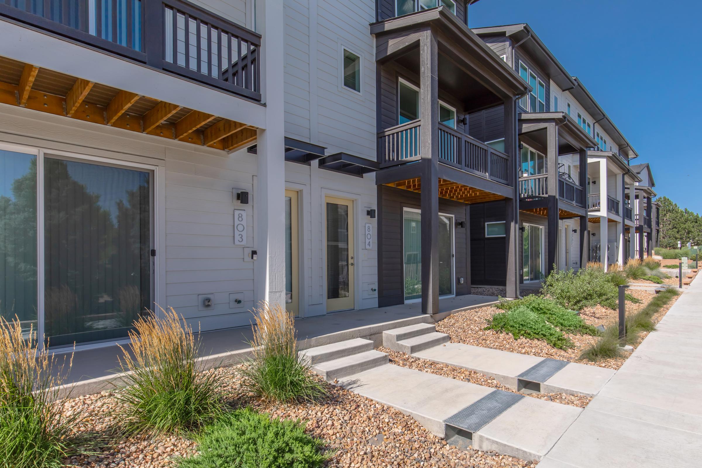 A row of modern townhouses with multiple levels, featuring wooden balconies and large windows. The ground is landscaped with rocks and grasses, and a concrete pathway leads to the entrances. The buildings have a combination of dark and light siding, set against a clear blue sky.