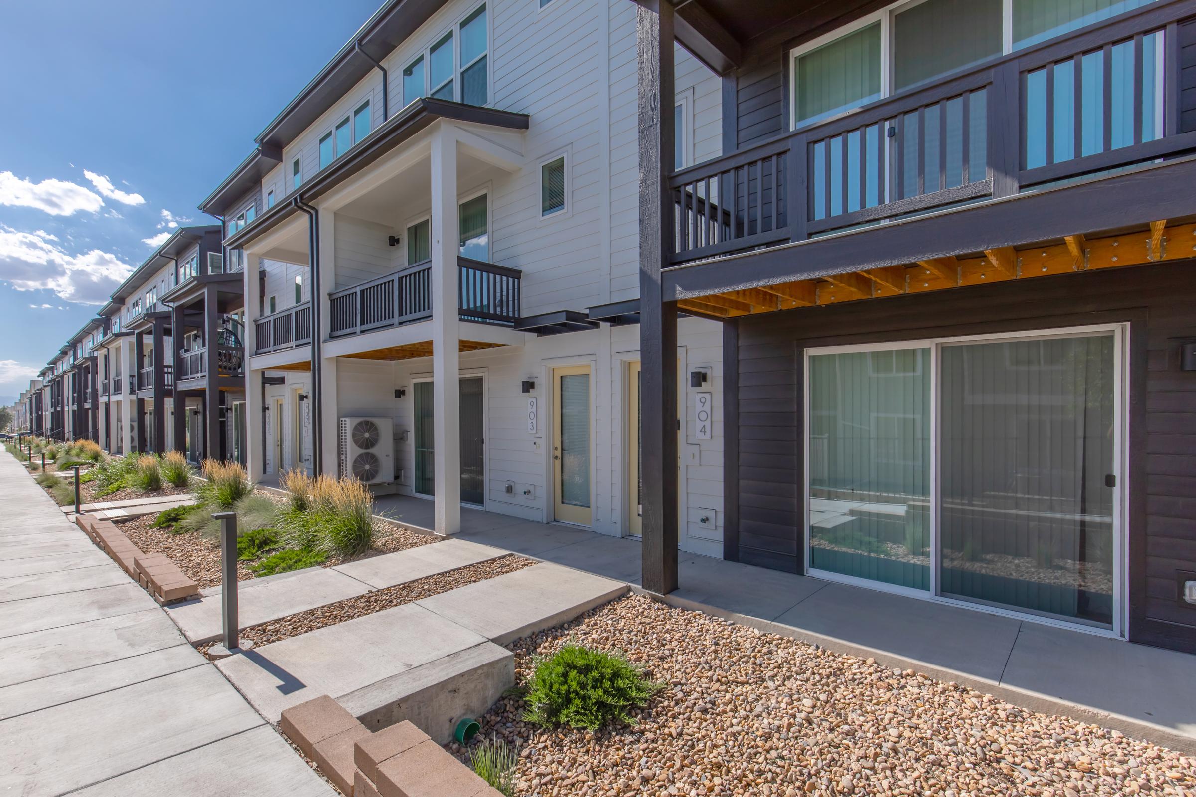 Row of modern townhouses featuring balconies, large windows, and concrete walkways. The architecture is sleek, with a mix of dark and light exterior colors. Sunlight is visible, with a clear blue sky in the background, and landscaped areas with plants and gravel enhance the curb appeal.