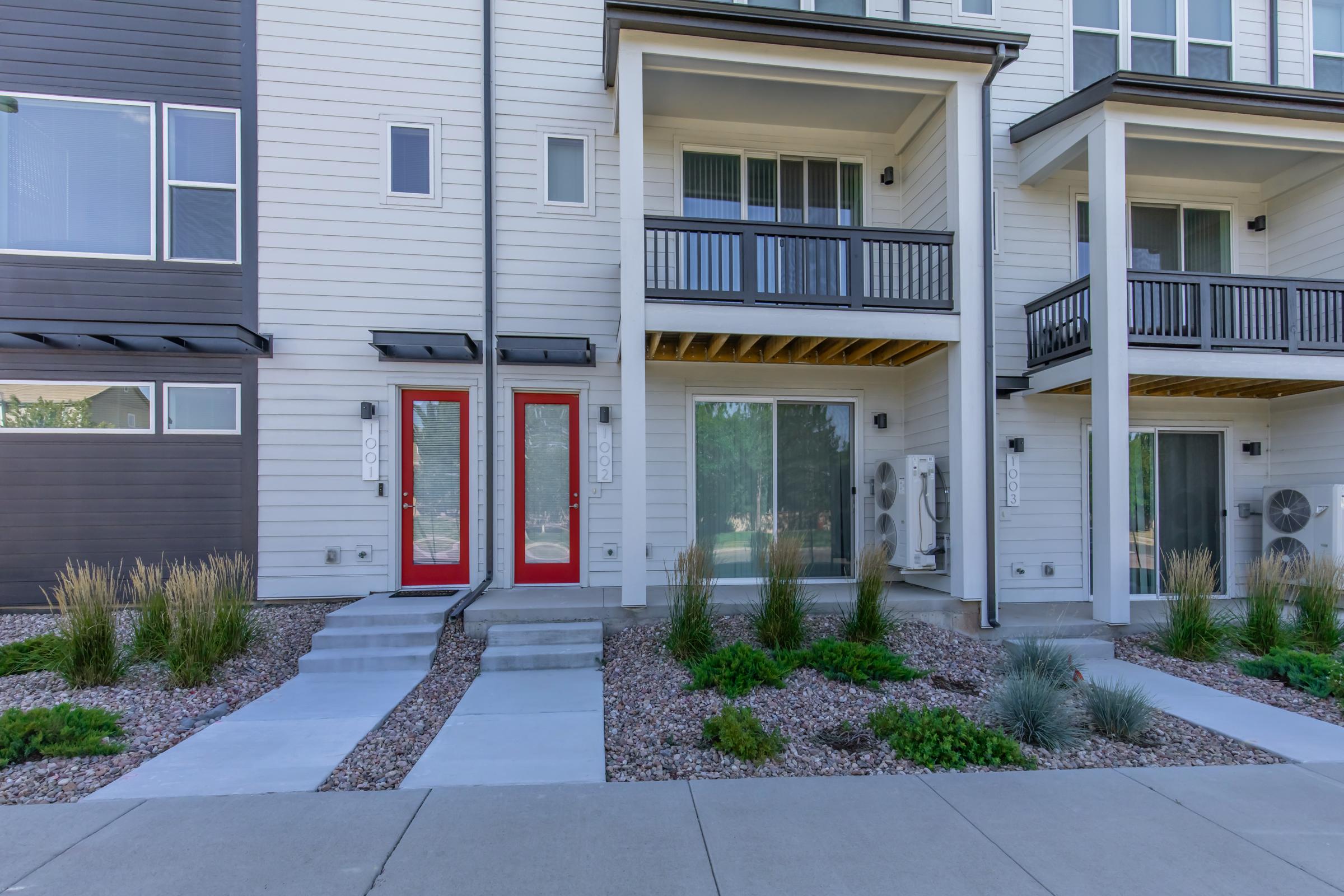 A row of modern townhouses with white and brown exteriors, featuring red front doors. The ground is landscaped with small shrubs and grasses, and there are concrete pathways leading to the entrances. Air conditioning units are visible beside the doors.