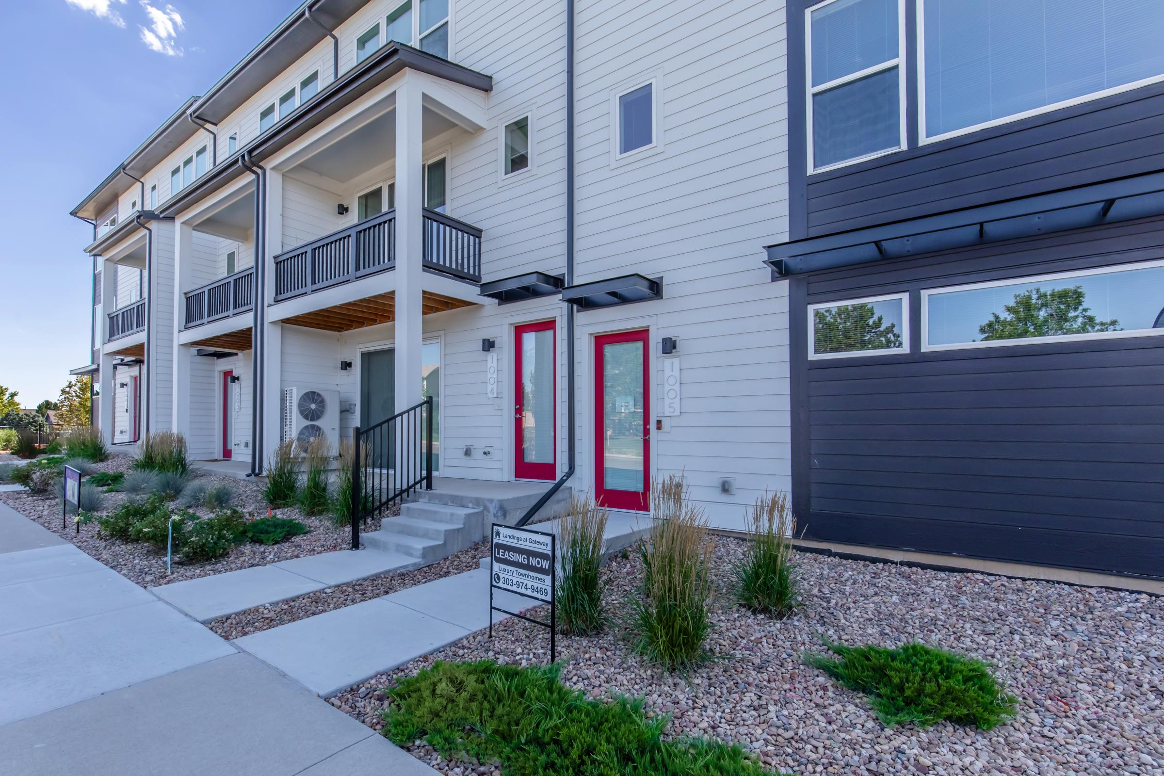 Exterior view of a modern townhouse community featuring two levels with multiple balconies, large windows, and red doors. The pathway leads to the entrance, surrounded by decorative landscaping including rocks and shrubs. A sign near the walkway indicates availability for leasing.