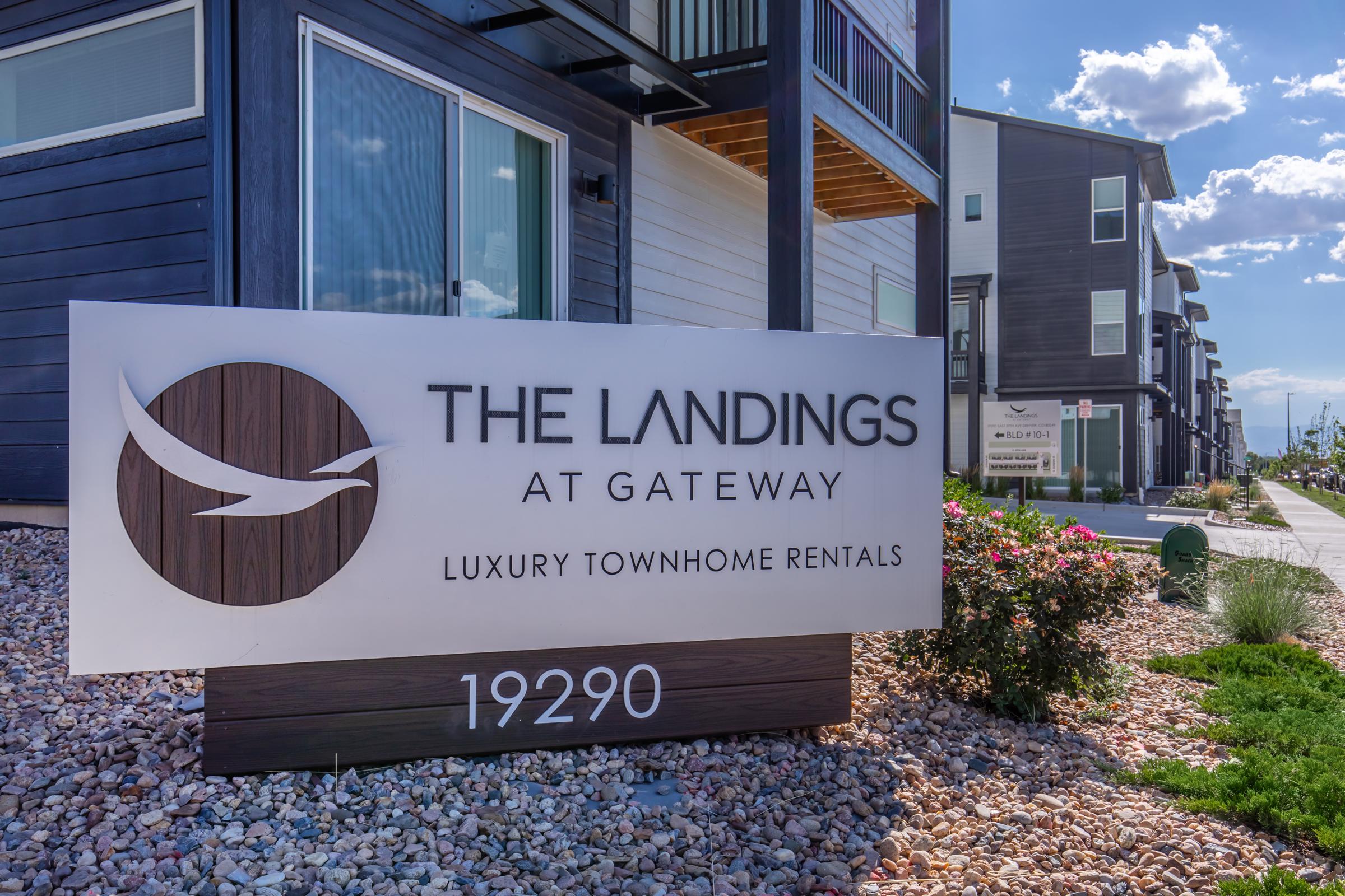 Sign for "The Landings at Gateway" featuring the text "Luxury Townhome Rentals" with a decorative logo. The sign is set against a backdrop of modern townhome buildings and landscaped areas, with blue skies and scattered clouds visible. The address "19290" is also displayed on the sign.
