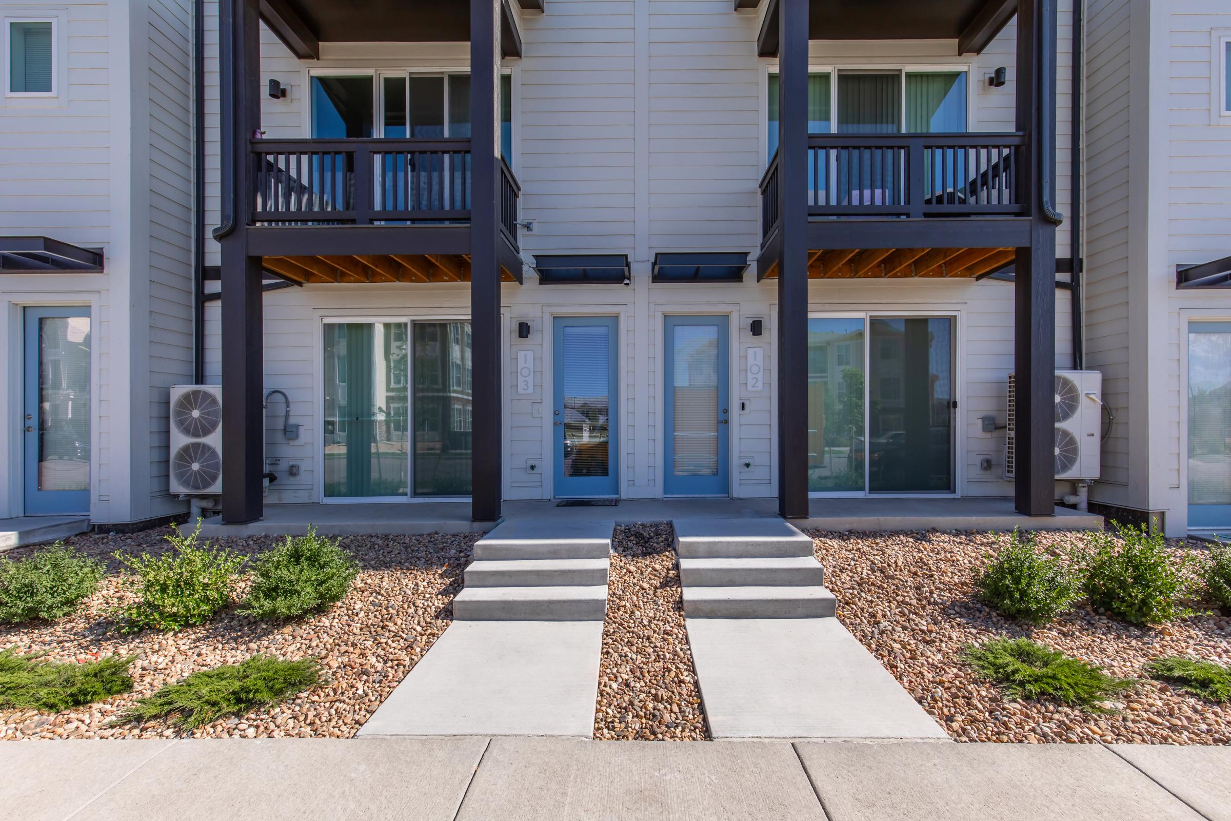 A modern townhouse exterior featuring two units with stairways leading to the front doors. The facade includes large windows, balconies, and air conditioning units, surrounded by landscaped gravel and small bushes. Bright sunlight highlights the clean, contemporary design.
