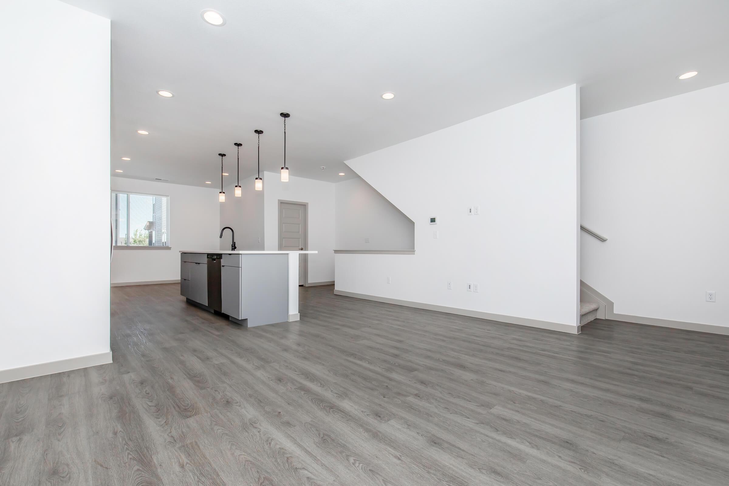 Modern, spacious interior of a newly constructed home featuring an open floor plan. The room has light gray laminate flooring, white walls, and a kitchen island with pendant lighting. There is a staircase in the background leading to an upper level, and large windows allowing natural light.