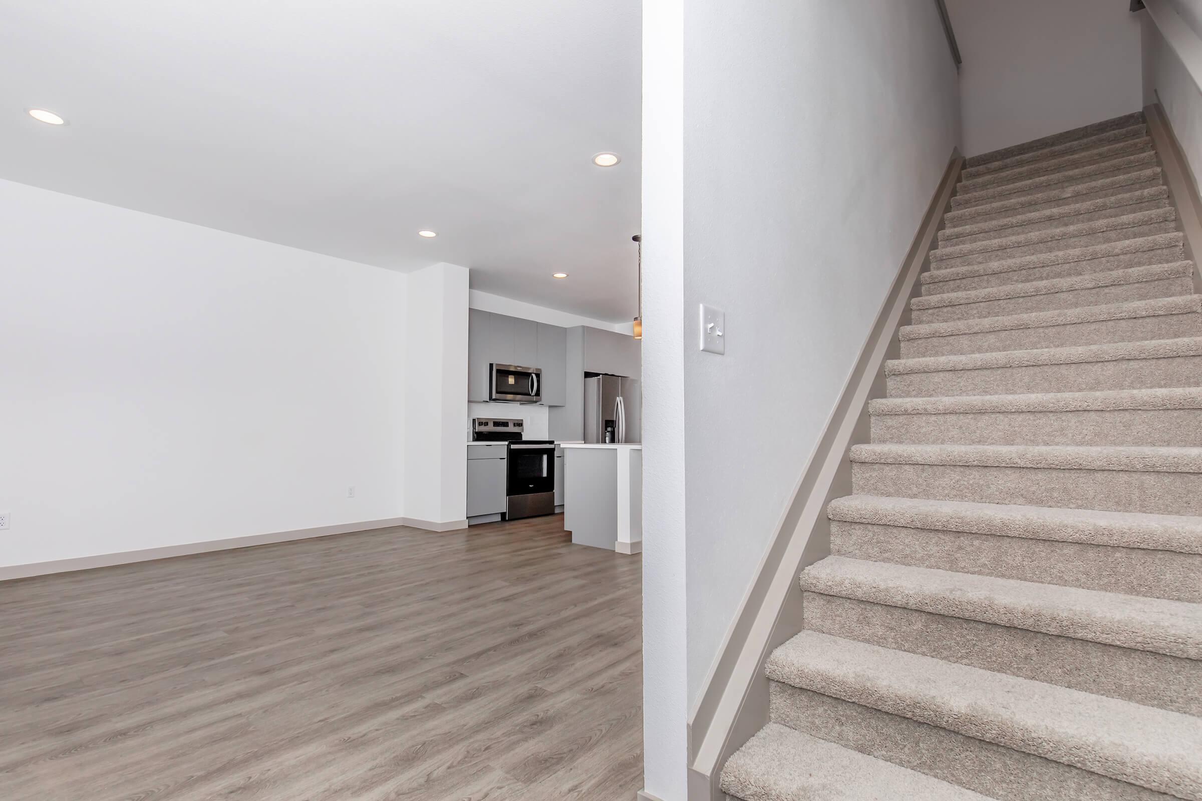 A spacious, modern living area featuring a light-colored floor, open kitchen with stainless steel appliances in the background, and a stairway leading to the upper floor with carpeted steps. The walls are painted a soft white, creating a bright and airy atmosphere.