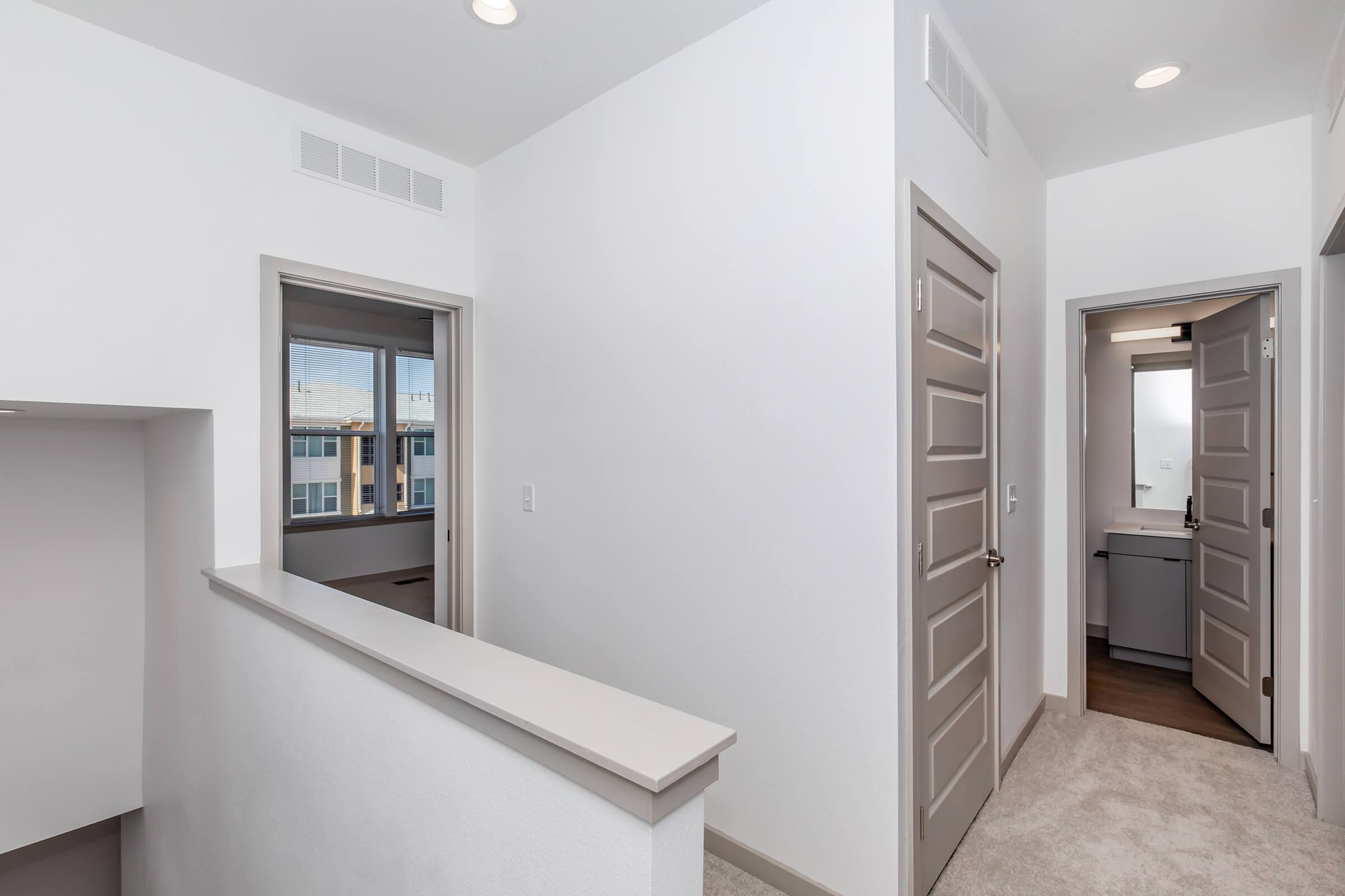 A well-lit hallway in a modern home featuring light gray walls, beige carpet, and two open doors leading to adjacent rooms. The space includes a railing overlooking a lower level and natural light streaming in from windows. The overall design is clean and contemporary.