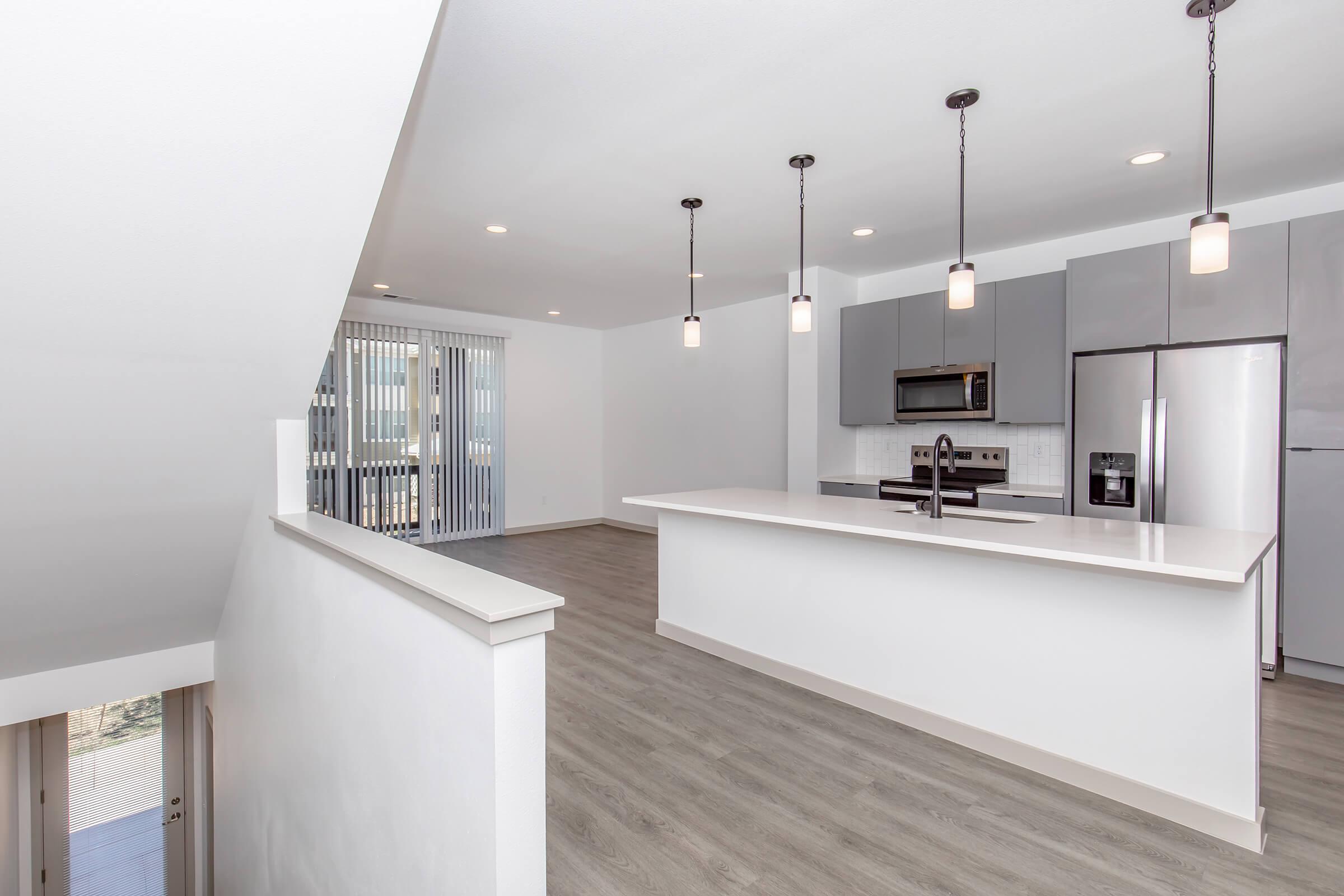 Modern kitchen and living space featuring gray cabinets, a large island with a dark faucet, pendant lighting, and stainless steel appliances. Bright open layout with wood-style flooring and large windows, allowing natural light to fill the room. Sliding glass doors lead to an outdoor balcony.
