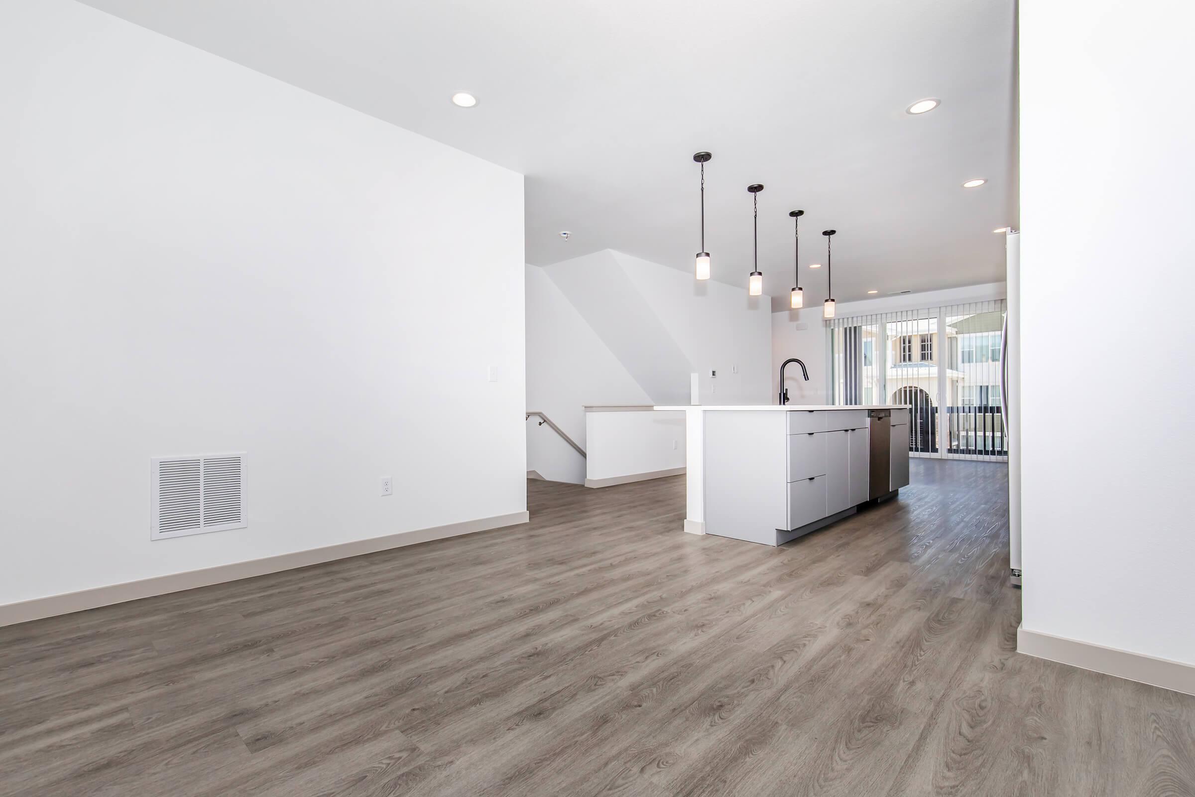 A modern, open-plan living space featuring light wood flooring, white walls, and a contemporary kitchen island with pendant lights. Large windows provide natural light, and sliding glass doors lead to a balcony. The layout offers a spacious feel with a stairway visible in the background.