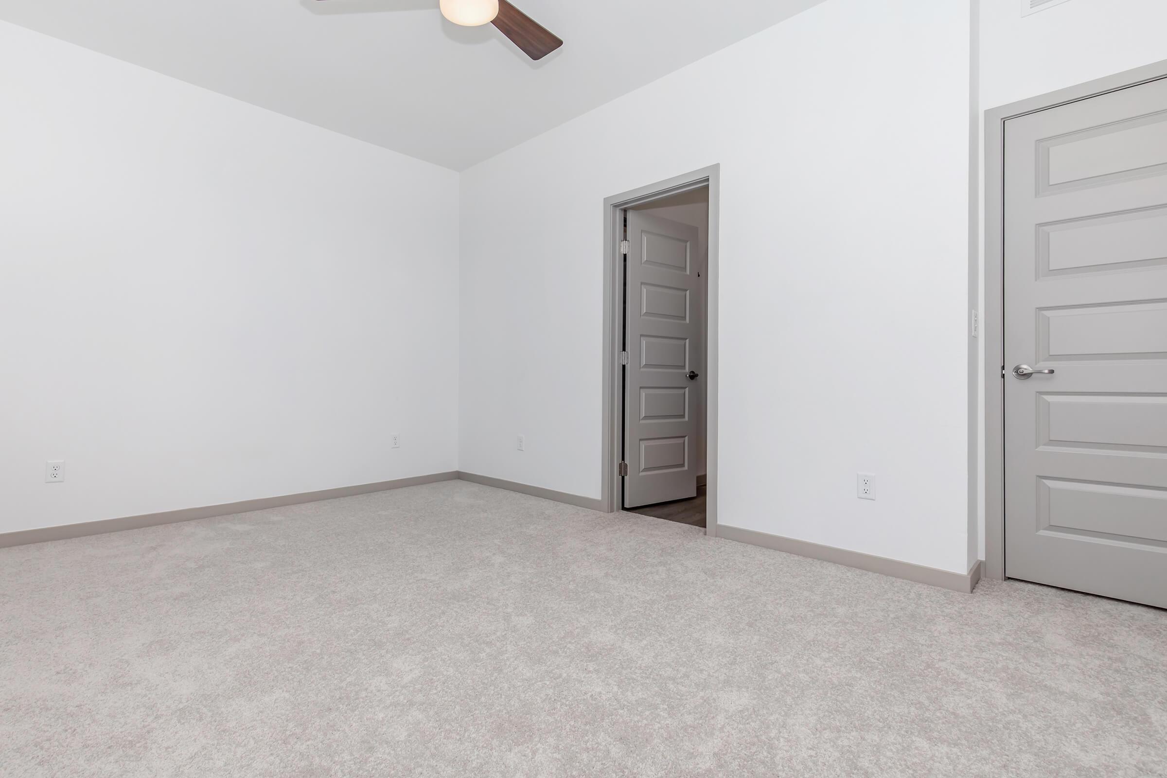 A minimalistic room with light gray walls and carpet. There is a ceiling fan and a closed door on the right leading to another space. The room is empty, offering a spacious feel, with natural light coming through an unseen window.