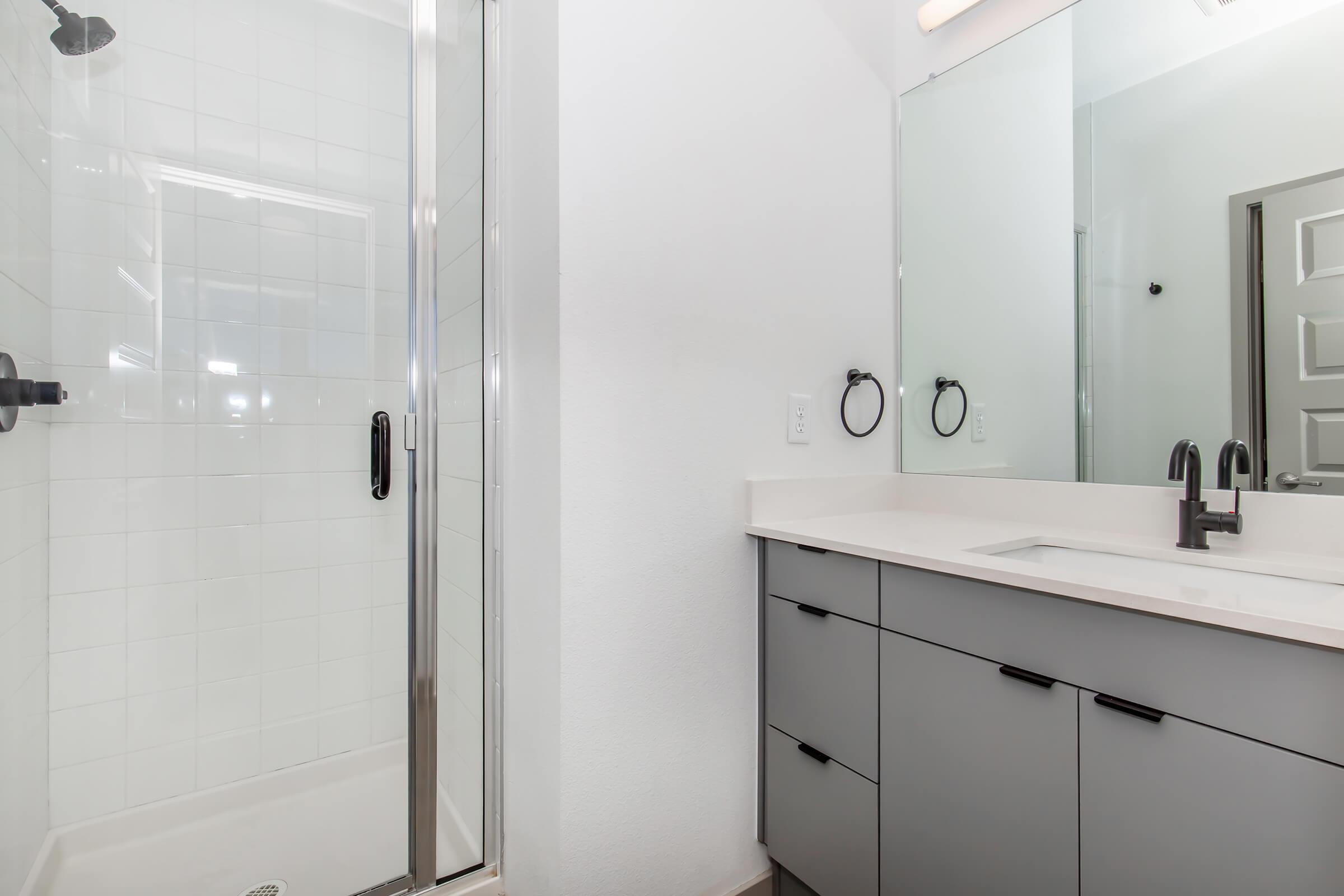 A modern bathroom featuring a glass shower stall with black fixtures, a light-colored vanity with drawers, and a large mirror above the sink. The space has neutral-toned walls and a sleek, minimalist design.