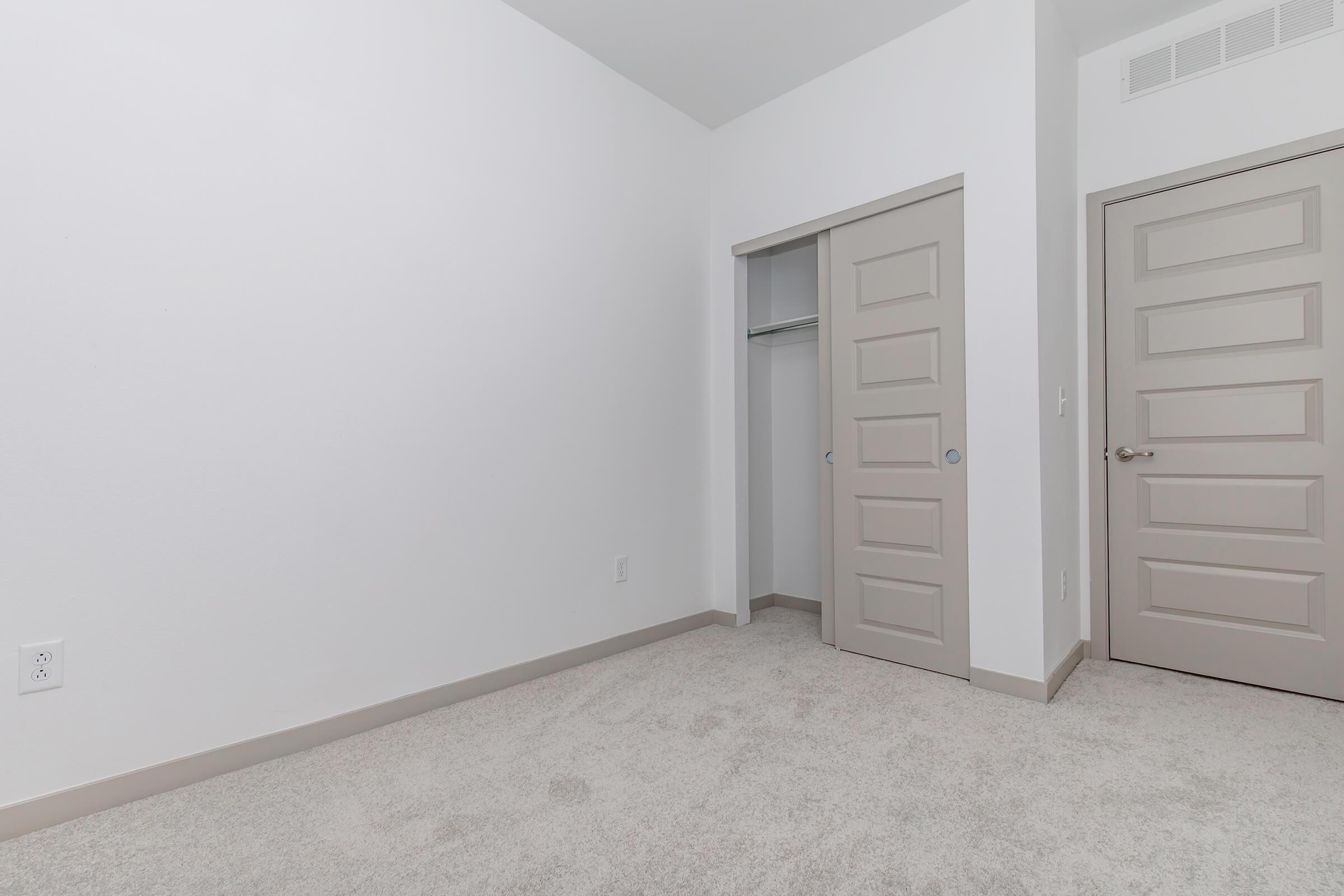A minimalist interior of a room featuring beige carpet flooring, white walls, and a closet with sliding doors. A door is visible on the right, both doors are a light, neutral color. The room appears empty and well-lit, with no furniture or decorations present.