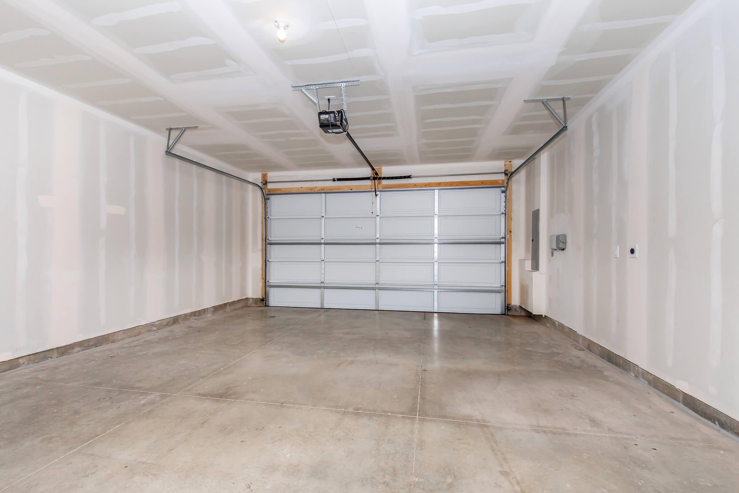 Empty garage with drywall walls and a concrete floor. The garage door is closed, and the ceiling is unpainted with visible light fixtures. There is a wall-mounted electrical panel on the side, and the space is well-lit and clean.