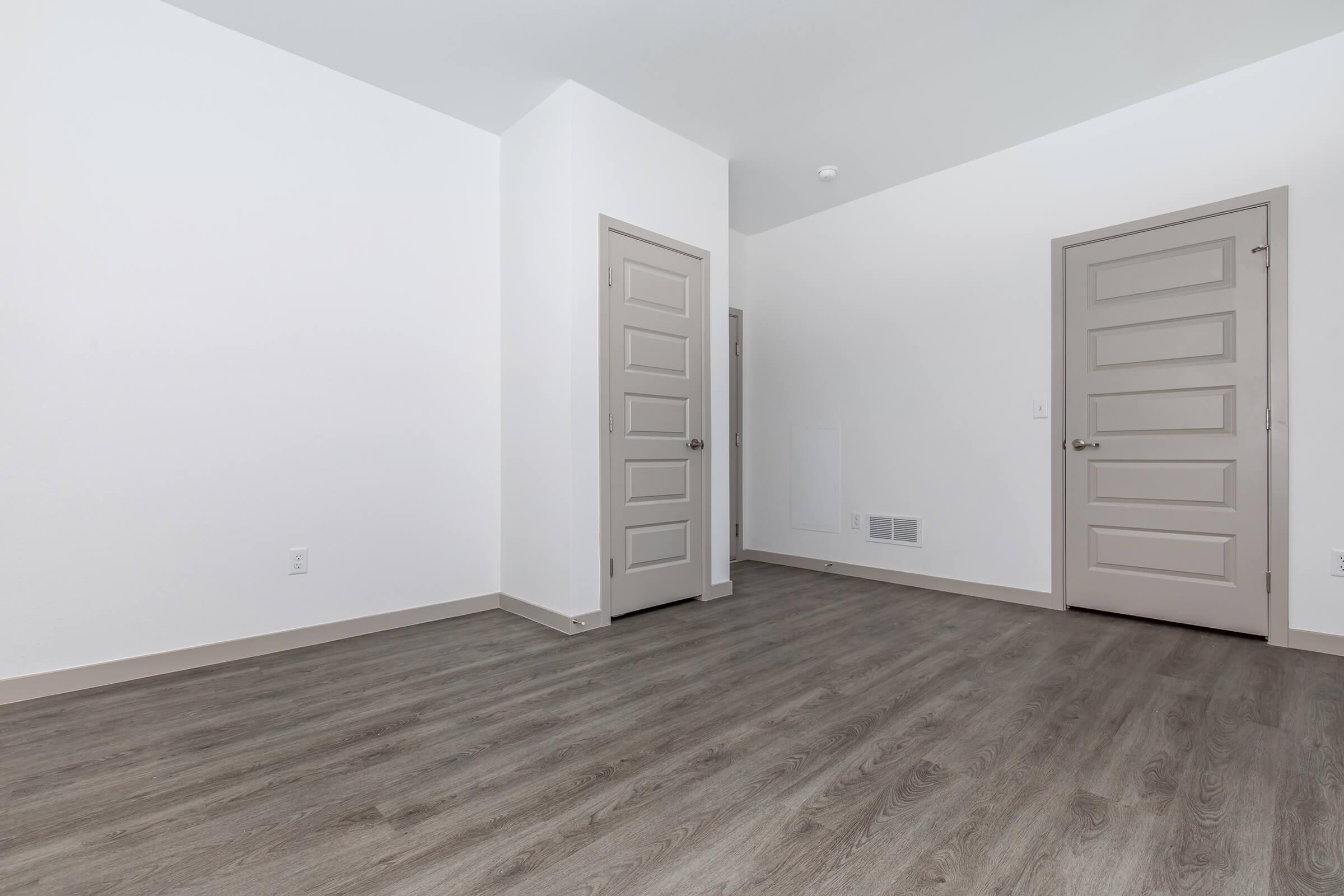 Empty room with light gray walls and a light-colored wooden floor. Two closed doors are visible on the right side, and the space is well-lit, creating a bright and open atmosphere. The room appears modern and freshly painted, with minimal furnishings.