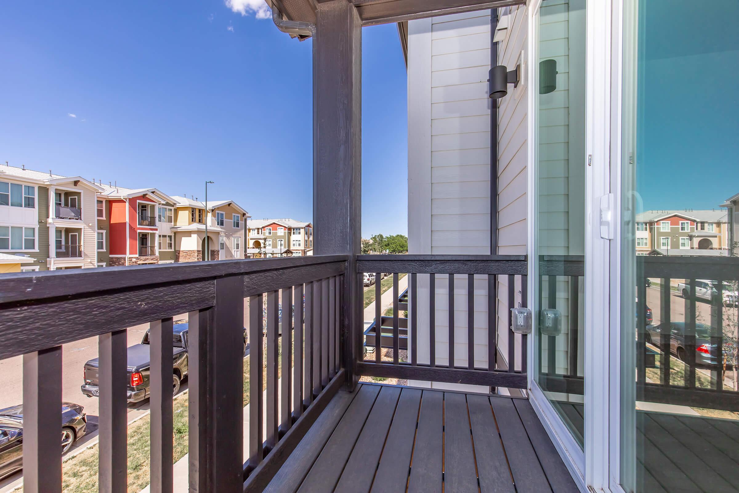 A view from a balcony featuring wooden railings and a floor made of planks. The scene shows neighboring buildings in various colors, with clear blue skies and a few cars parked below. The balcony has a glass sliding door that reflects part of the building.
