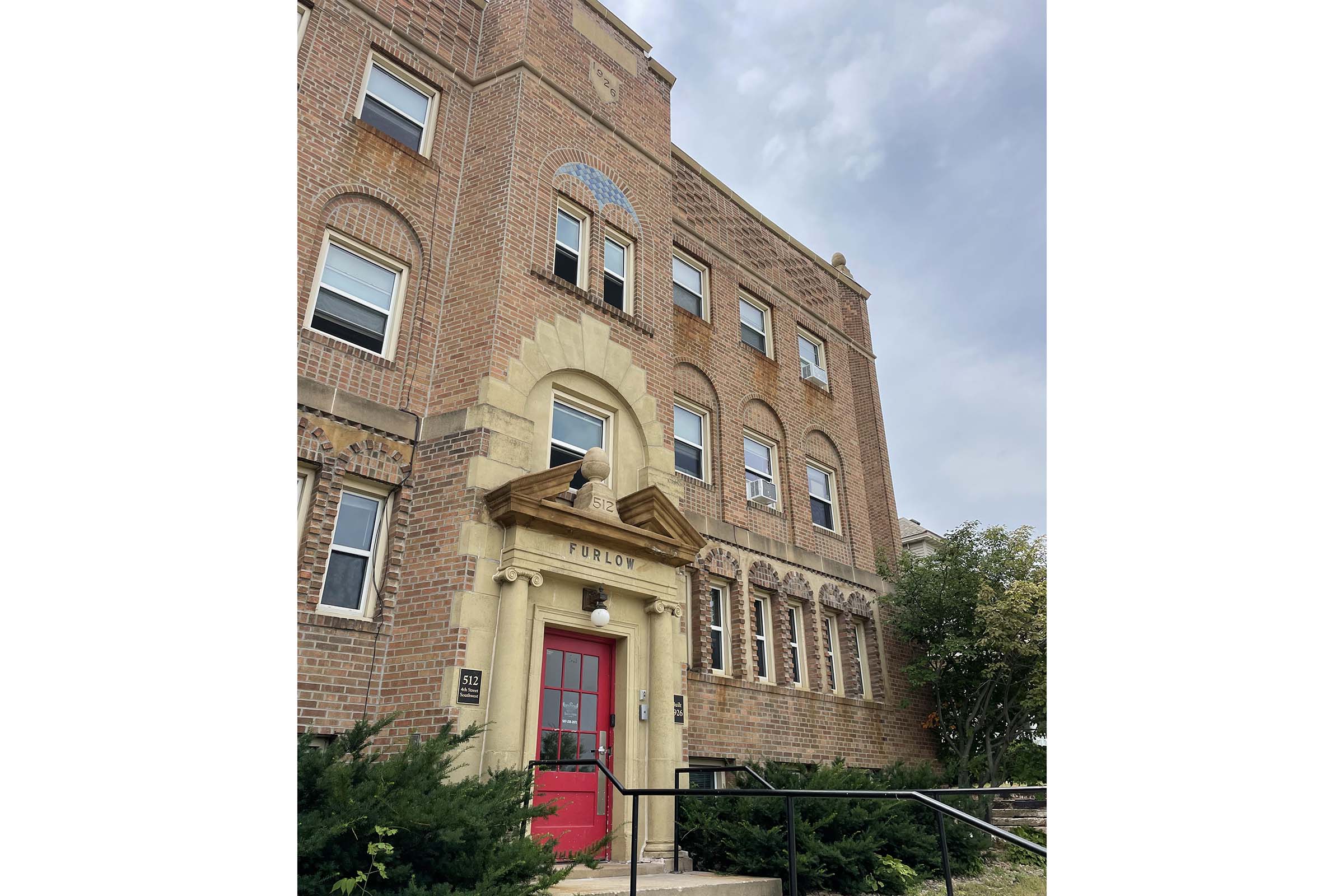 Brick building with a prominent entrance featuring decorative stonework above the door. The facade includes multiple windows, some with decorative arches. Lush greenery surrounds the base, and the sky is overcast, suggesting a cloudy day.