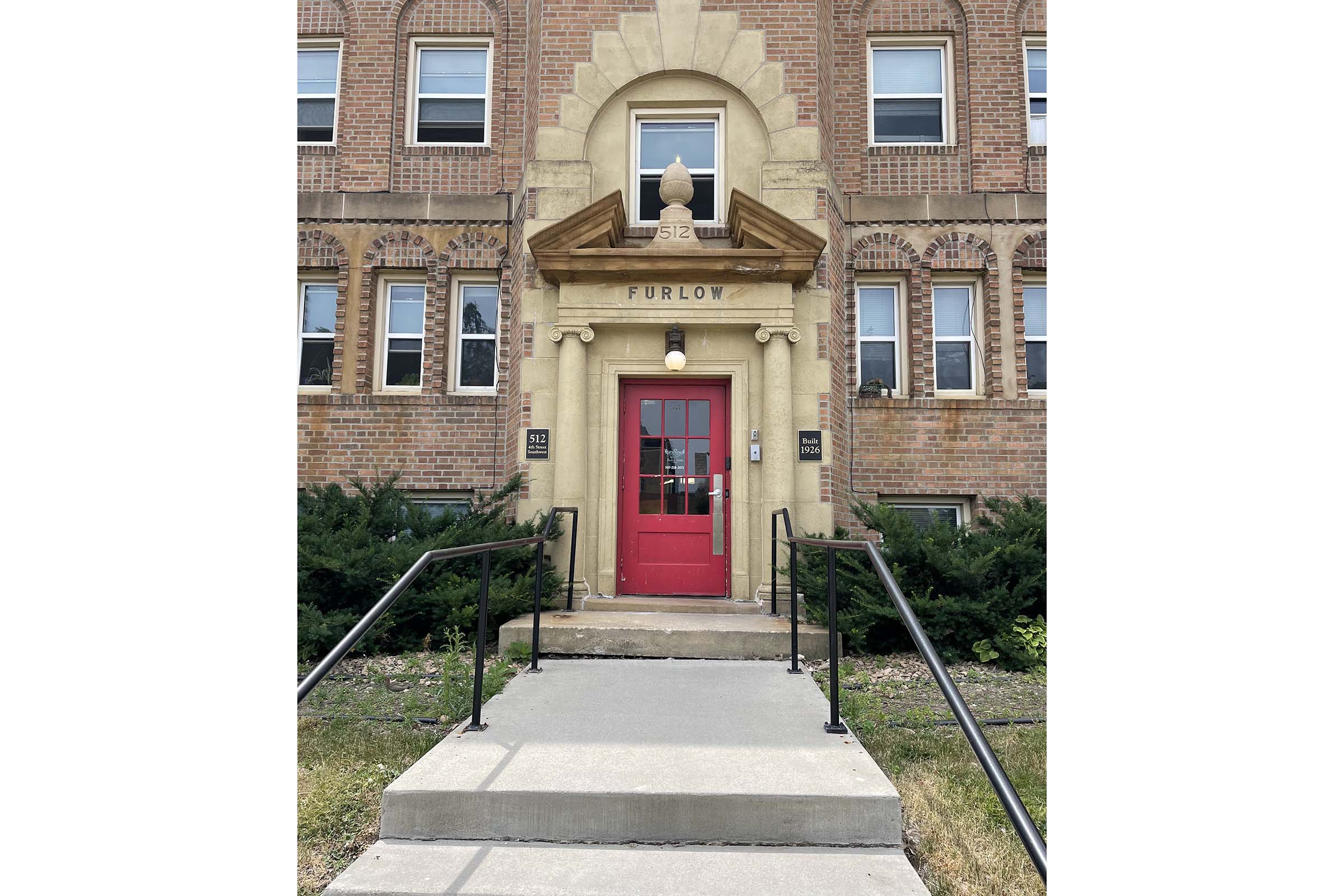Front entrance of a brick building with a prominent red door and decorative stonework above. The door features a white light fixture and is framed by a small overhang. The building has several windows and bushes lining the walkway, leading up to the entrance steps. The name "FURLOW" is visible above the door.